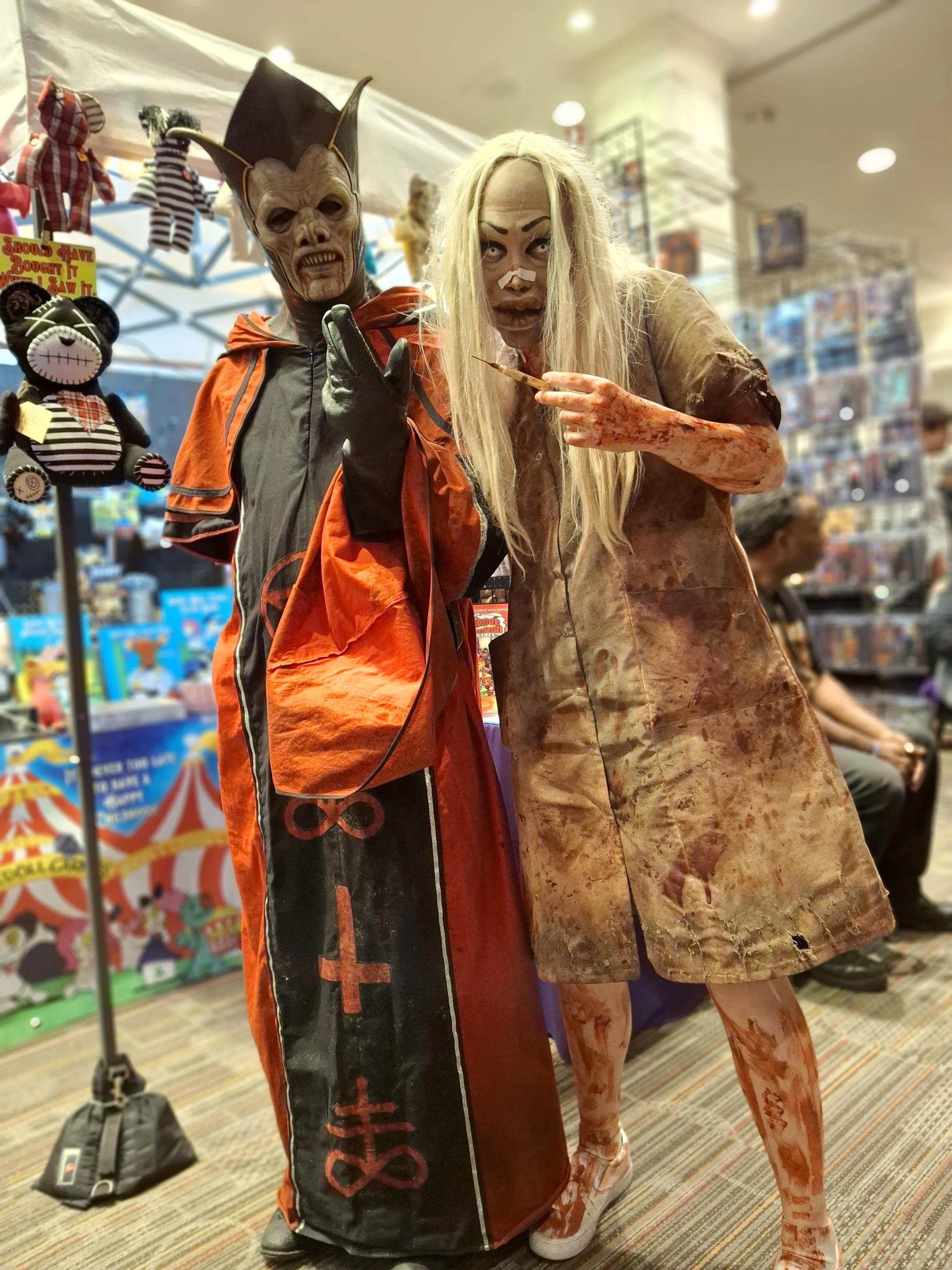 Two people in detailed horror costumes. One dressed as a demon with large horns, a mask with exposed teeth, and a red and black robe. The other as a bloody, deranged nurse with long blonde hair, bloodstained clothing, and holding a syringe. They are 
