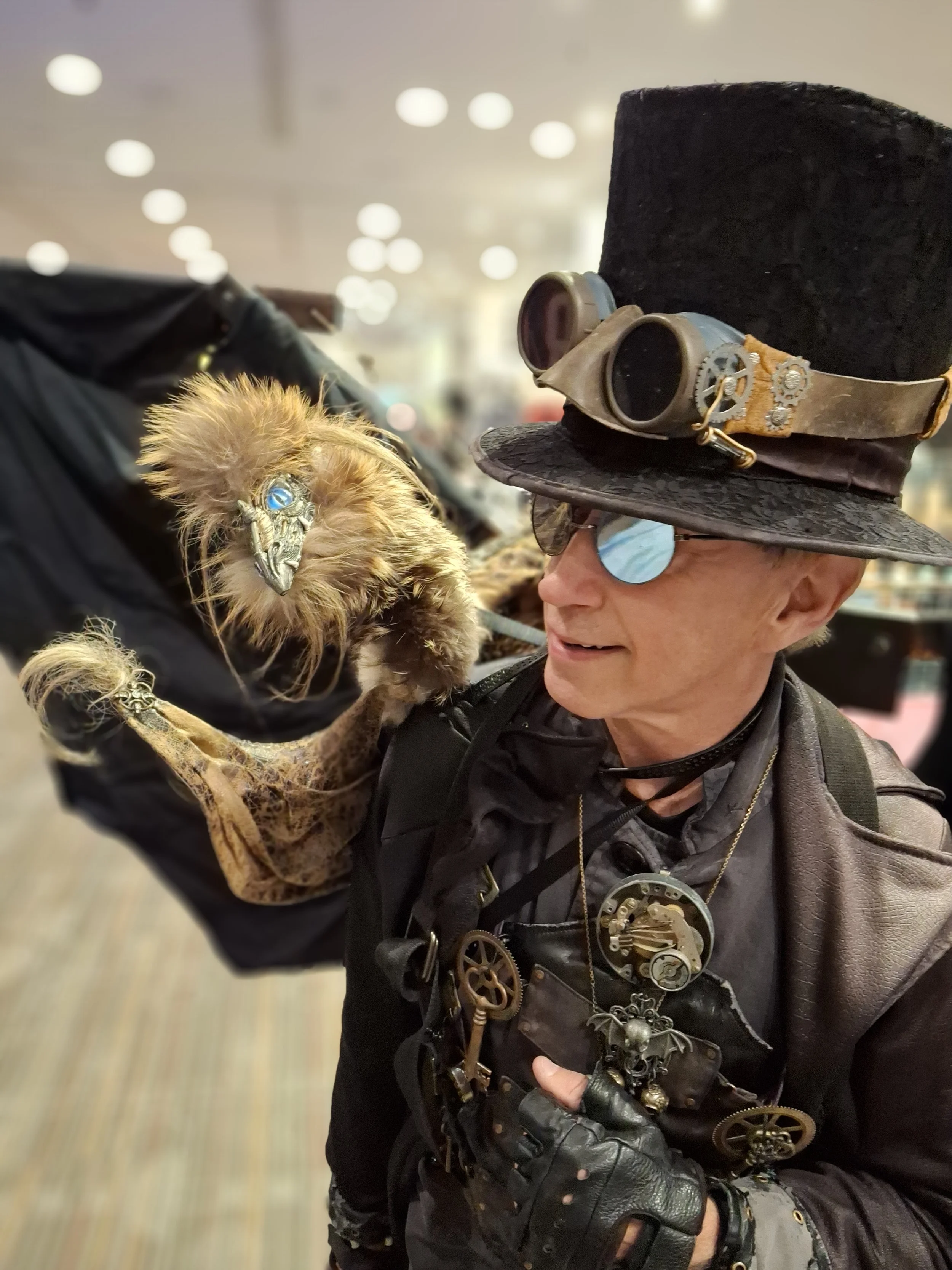 A person dressed in steampunk attire holding a plush bird with a metallic beak and blue eyes in a shopping mall.