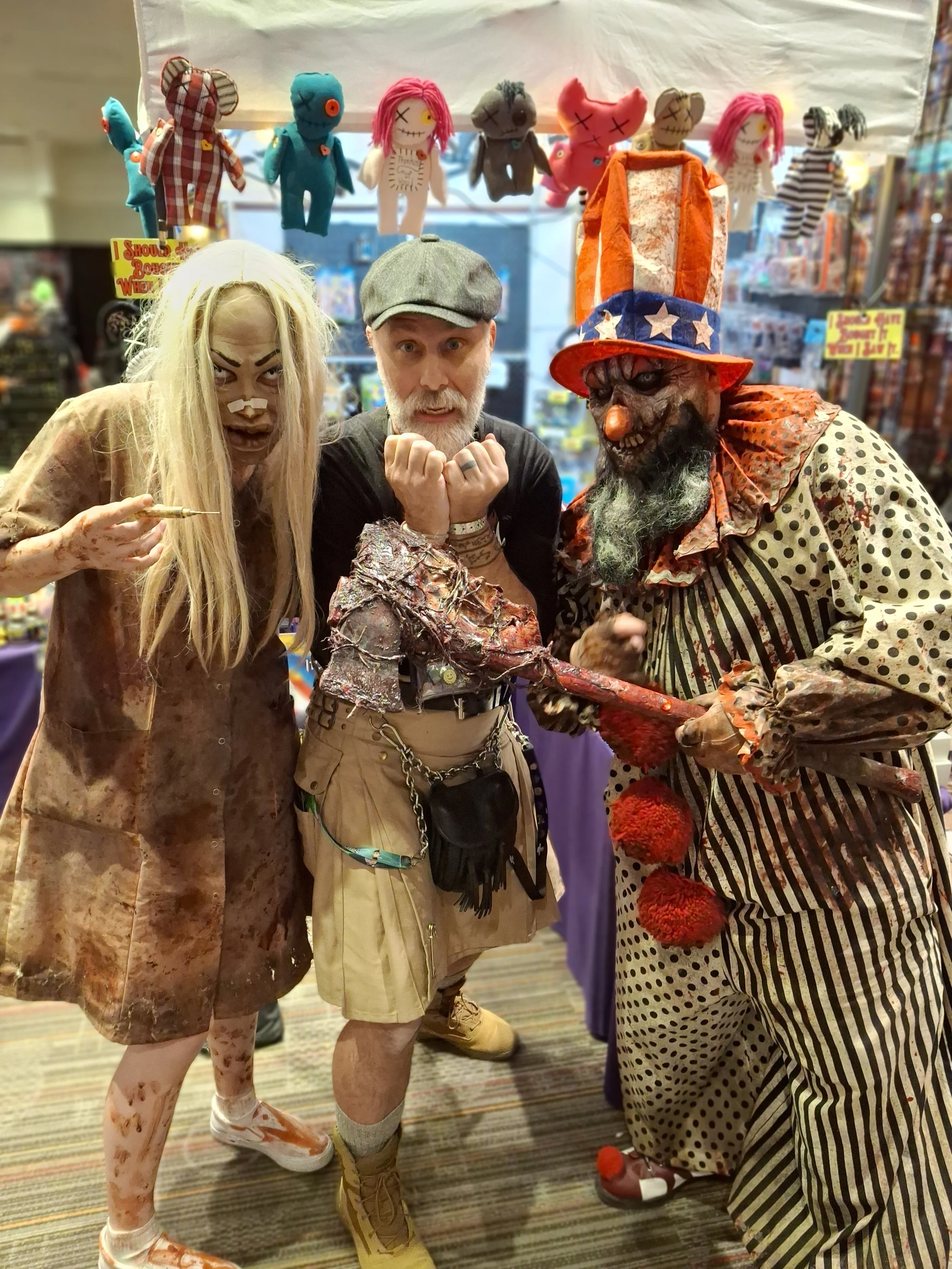 Three people dressed in Halloween costumes posing together at a store. One is dressed as a bloody, zombie-like figure with a knife; the second is dressed as a creepy clown with a large hat and a bearded face; the third is a person in casual clothing 