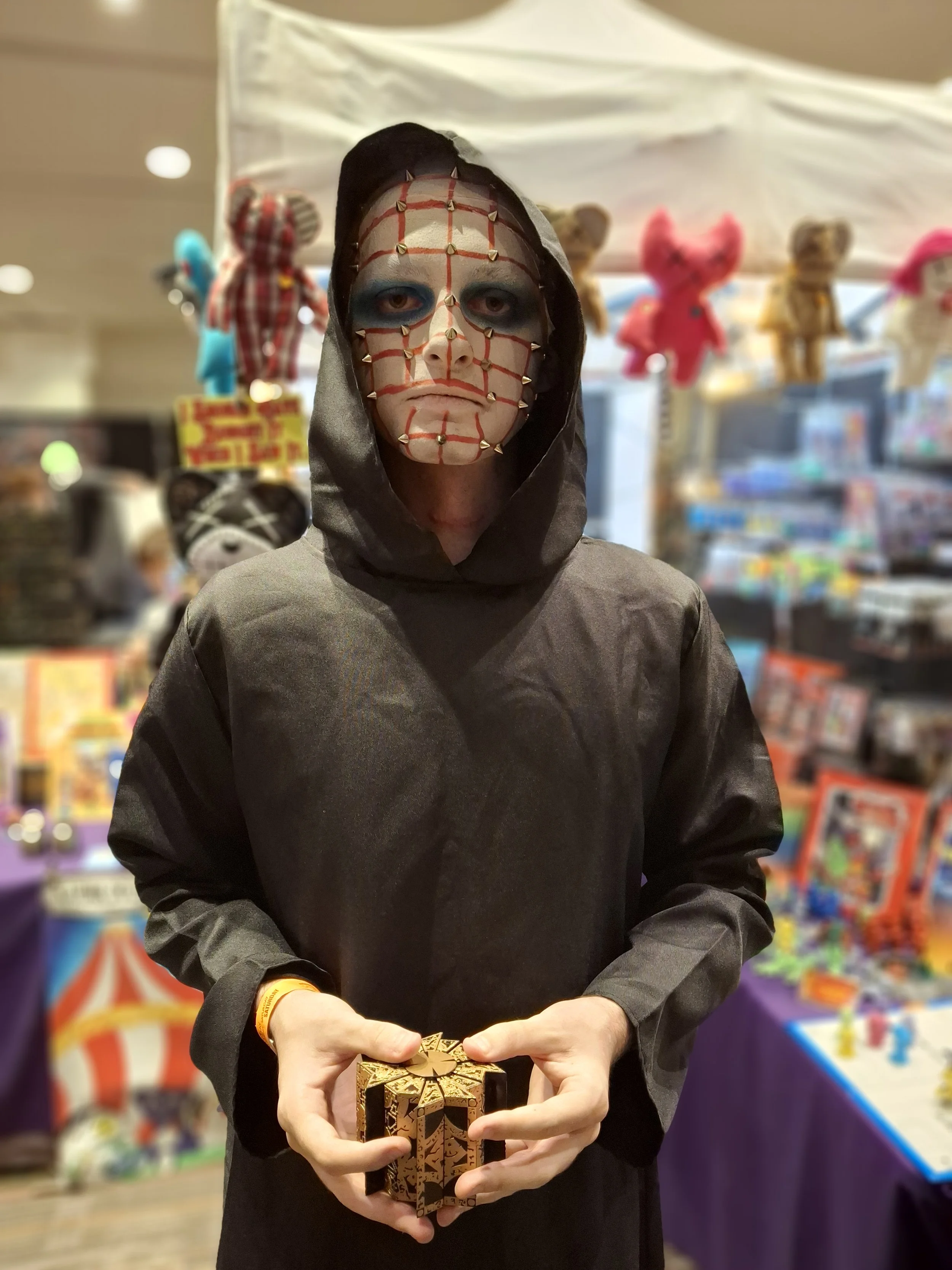 Person wearing a black hooded cloak and a mask with red strings and metal spikes, holding a gold and black patterned rectangular box, at a booth with toys and plush animals in the background.