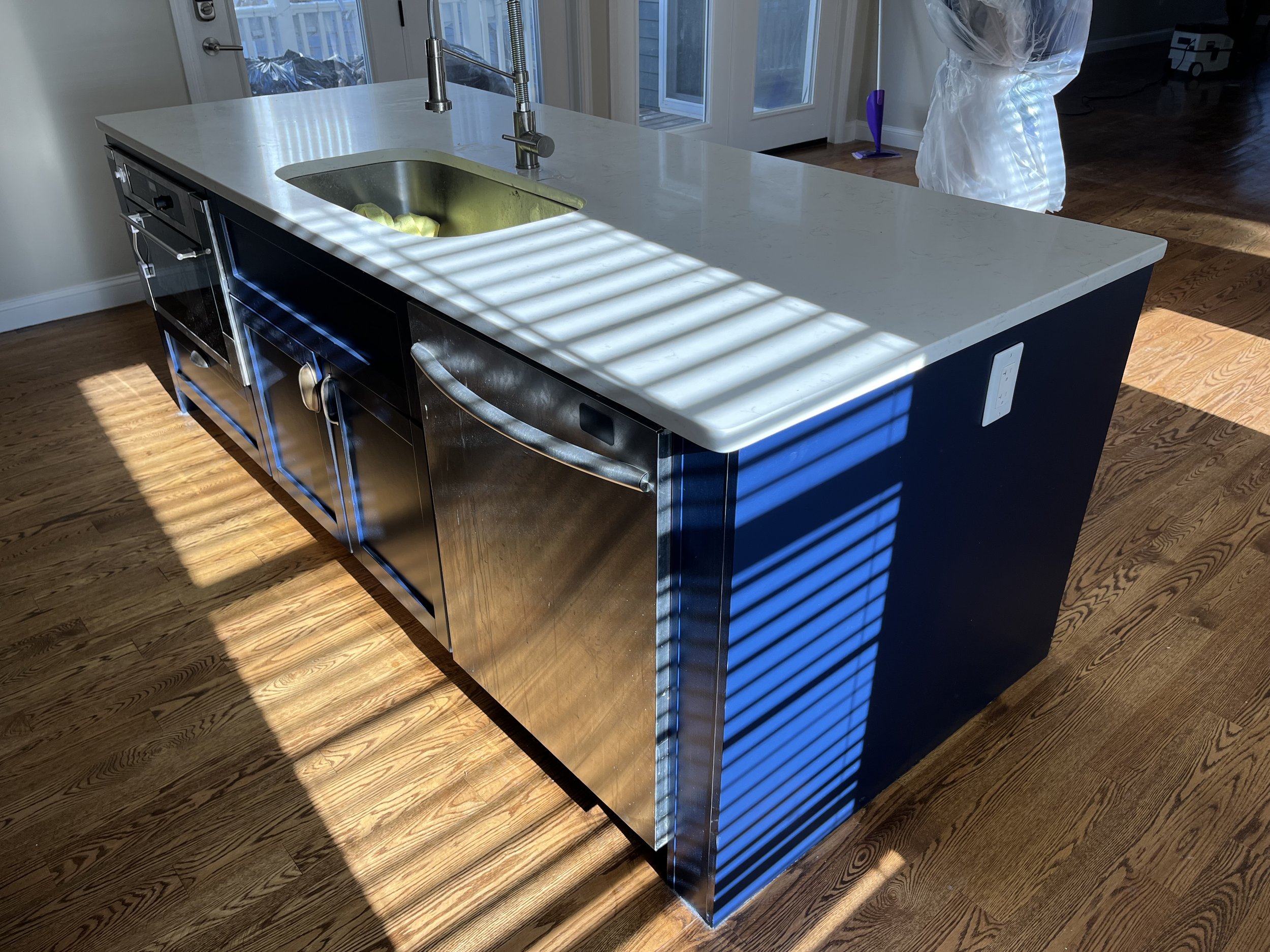 Modern kitchen island with white countertop, stainless steel sink, dishwasher, and cabinetry in a sunlit room.
