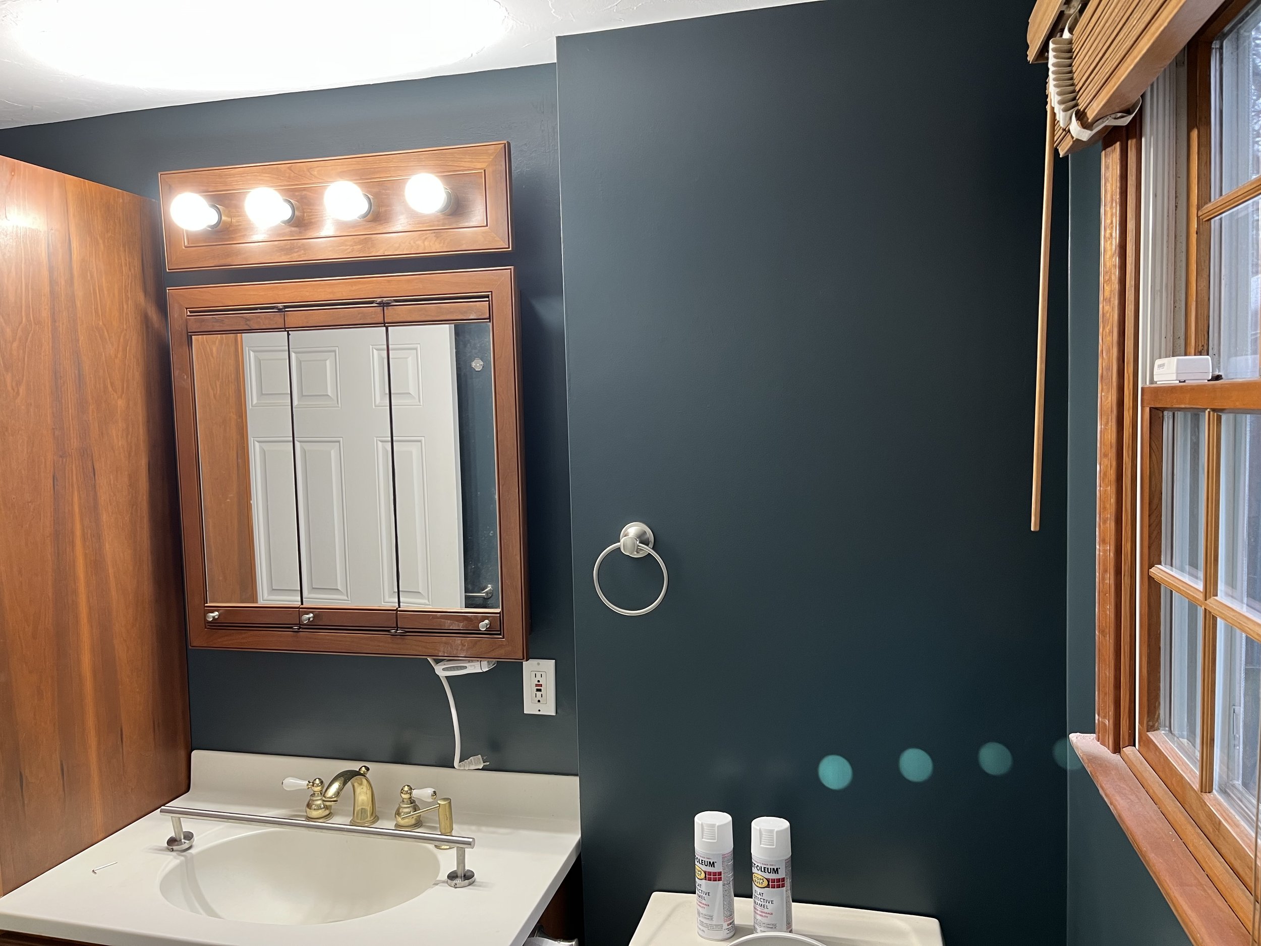 Bathroom interior with a dark green wall, wooden-framed mirror, vanity with white countertop, gold faucet, overhead lights, towel ring, window with blinds, and two cans on the counter.