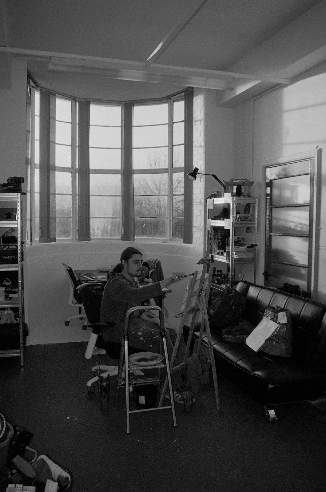 A person painting on a canvas in an art studio with large windows, bookshelves, a black sofa, and art supplies.