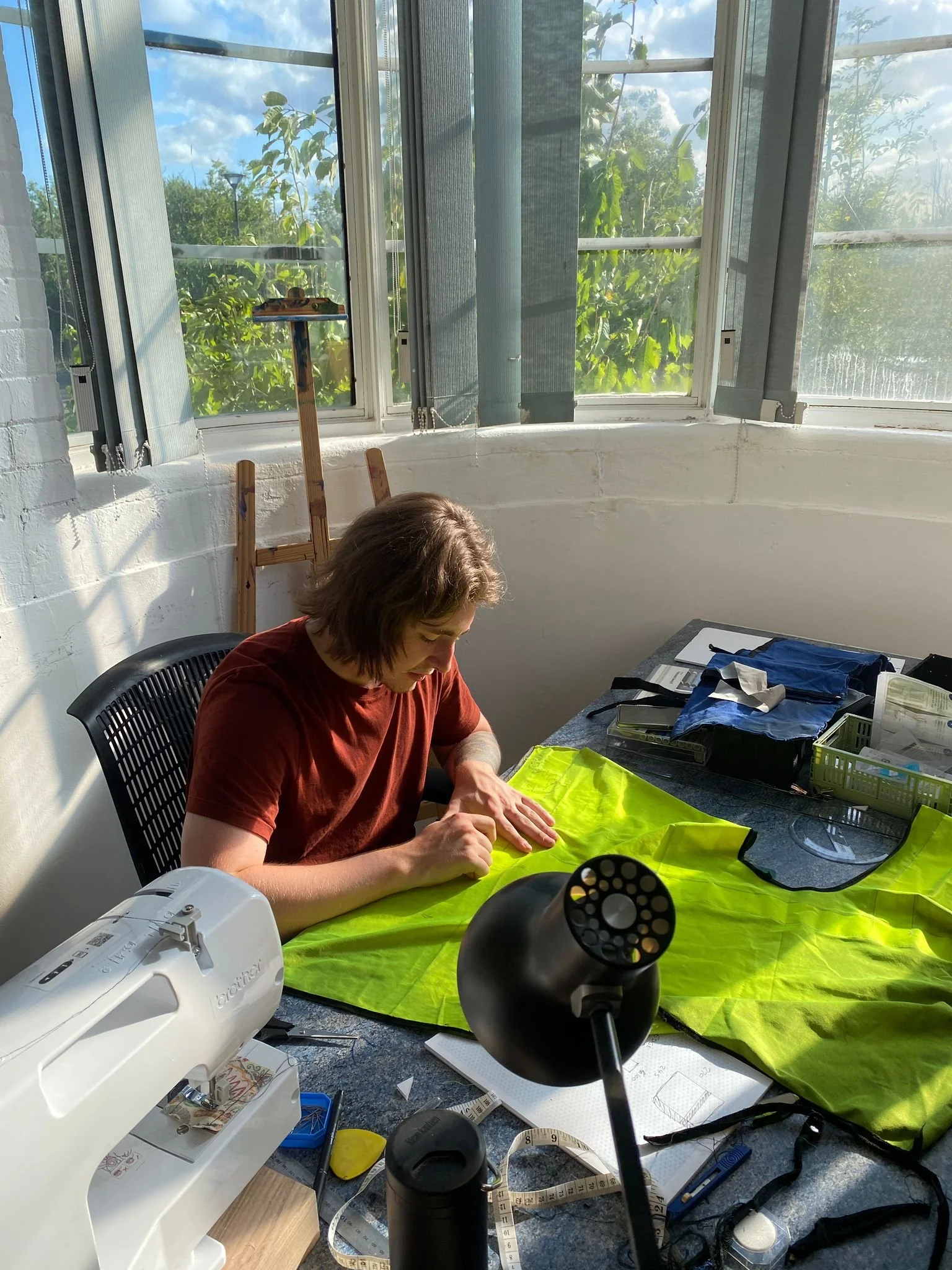 A person with brown hair wearing a red shirt is working on a project at a cluttered table inside a room. The scene is illuminated by sunlight coming through large windows with open blinds, showing trees outside. The table is covered with various sewi