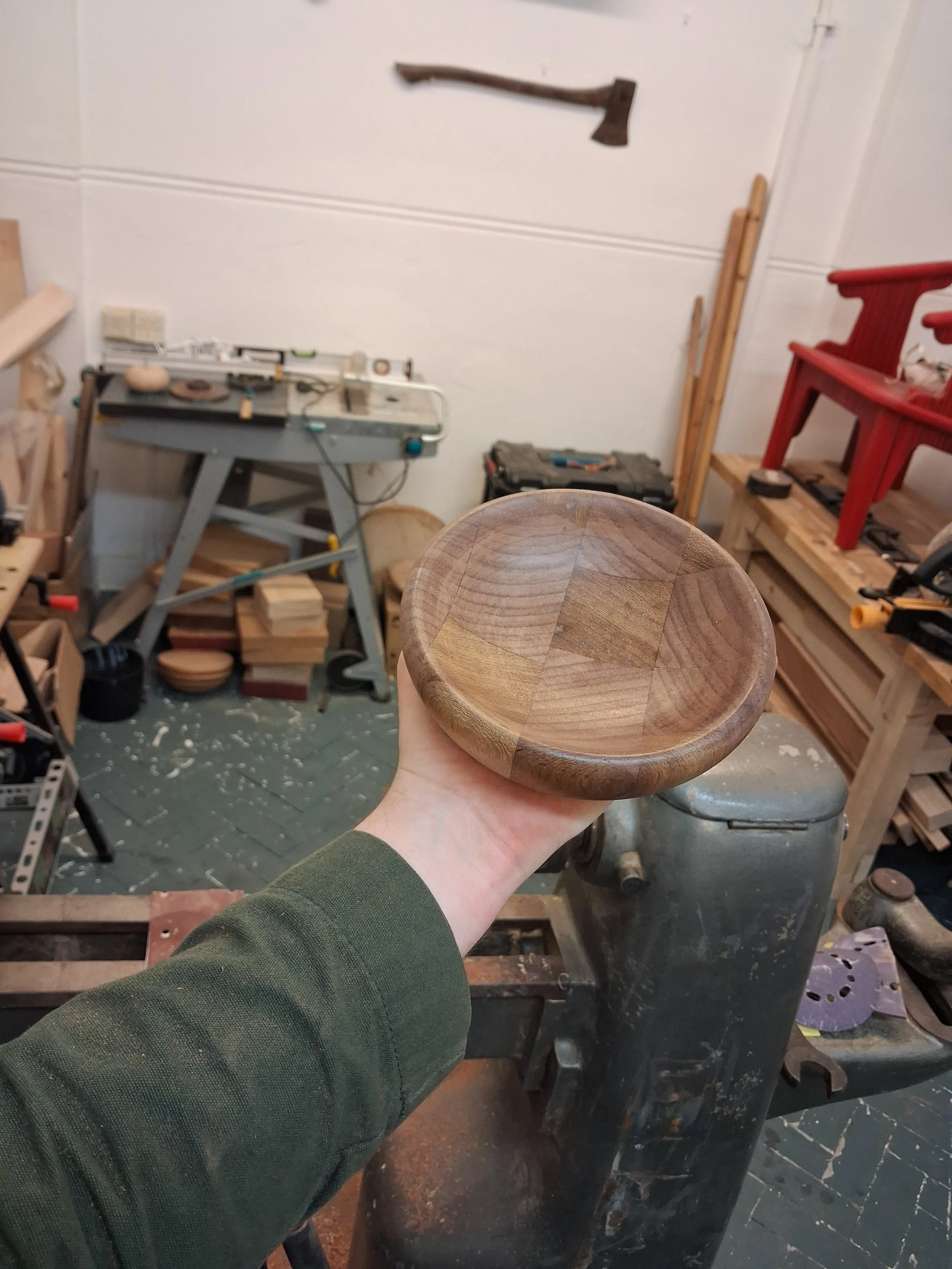 A person holding a wooden bowl in a woodworking workshop with tools, wooden pieces, and workbenches in the background.