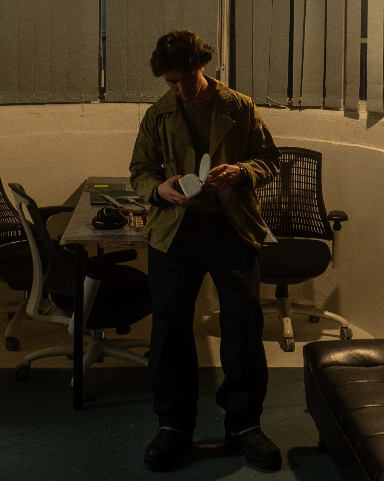 A person with short curly hair wearing a brown jacket and black pants standing in a dimly lit office or studio, holding a small white object, with office chairs, a desk, and various items in the background.