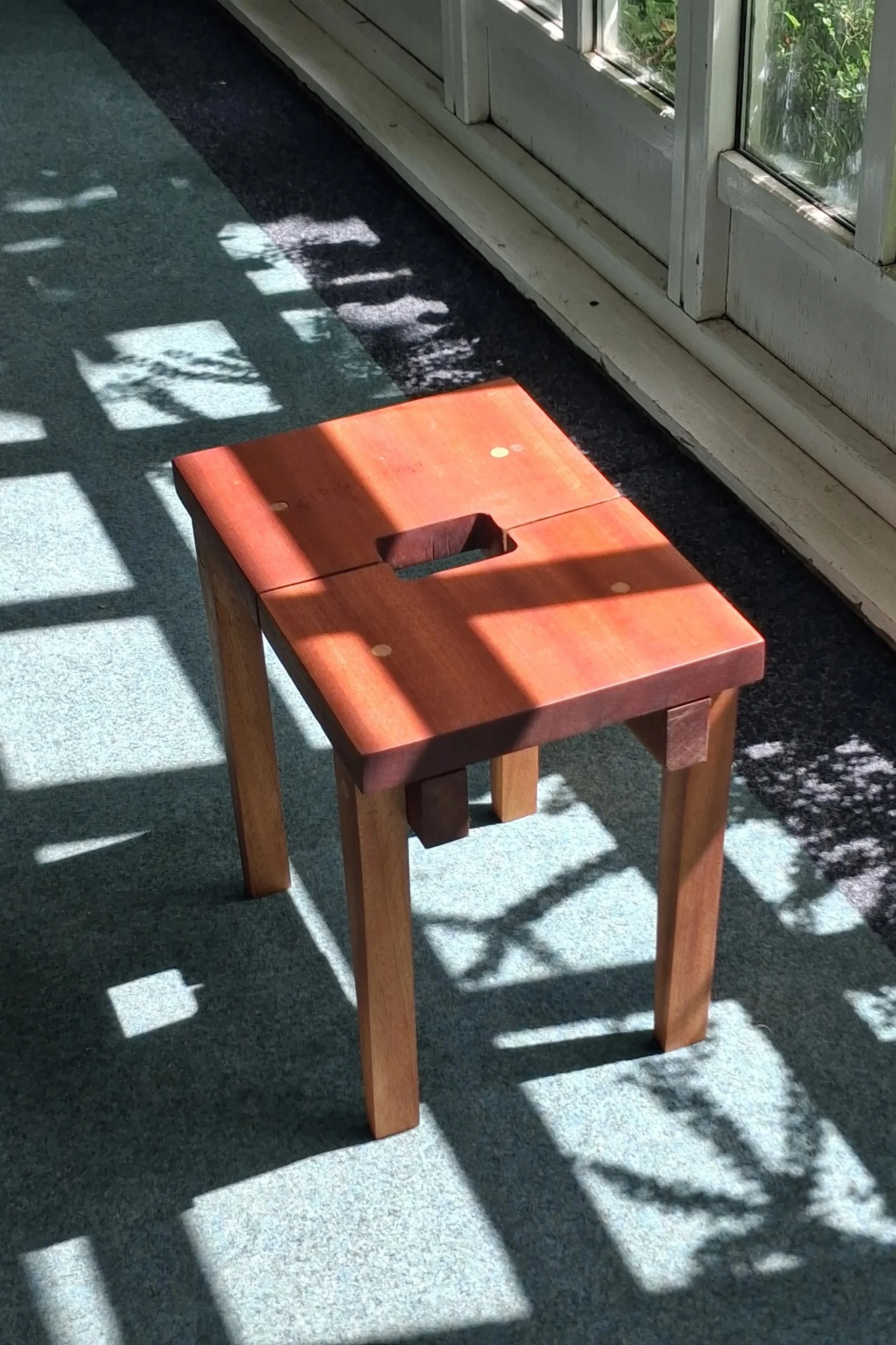 A small wooden stool with a square hole in the center, placed on a carpeted floor near a glass door. Sunlight creates shadows of the door's grid pattern on the floor and the stool.