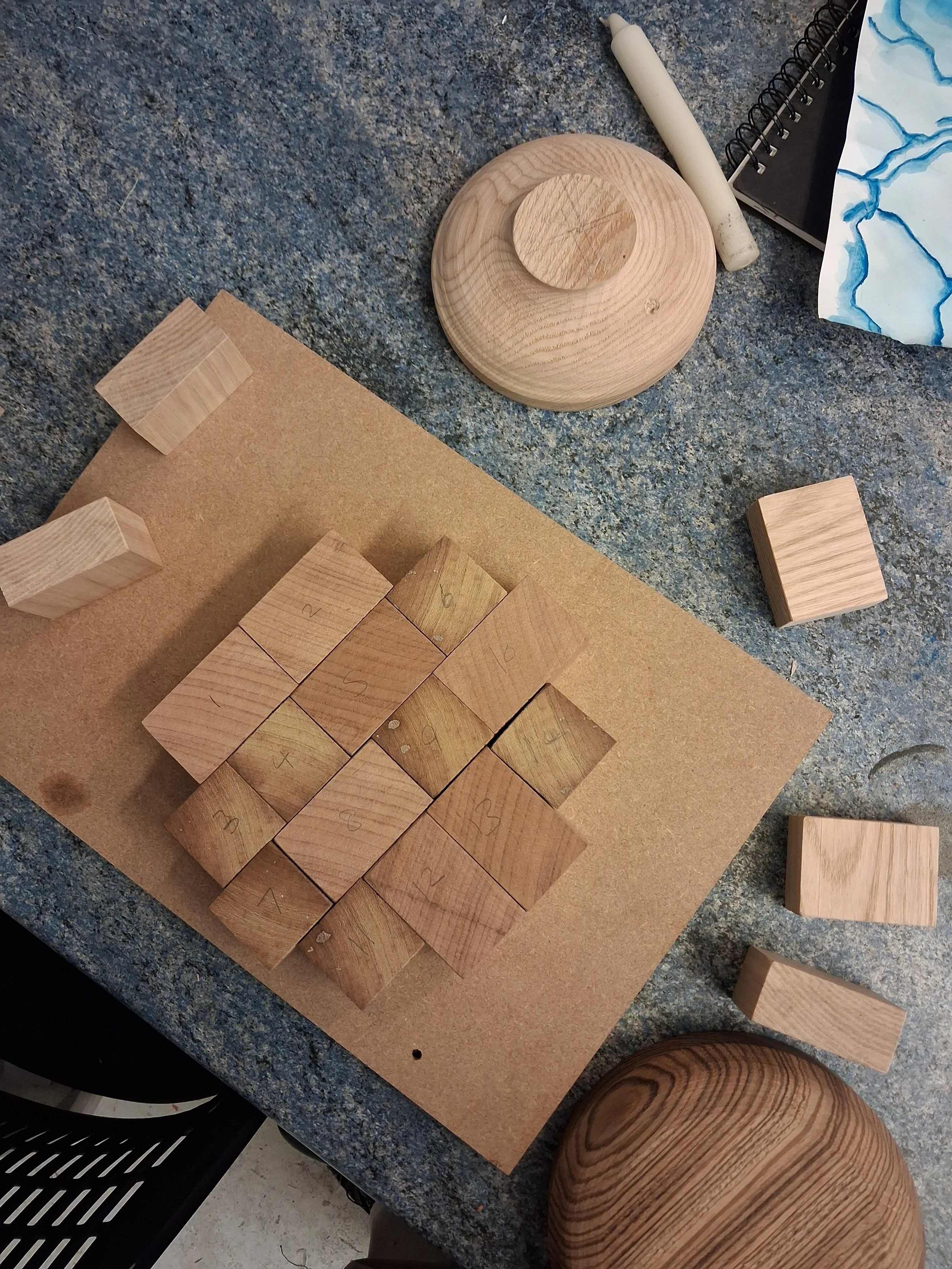 Wooden blocks and plates arranged on a speckled blue surface, some labeled with numbers and letters, with a spiral notebook and a white cylindrical object nearby.