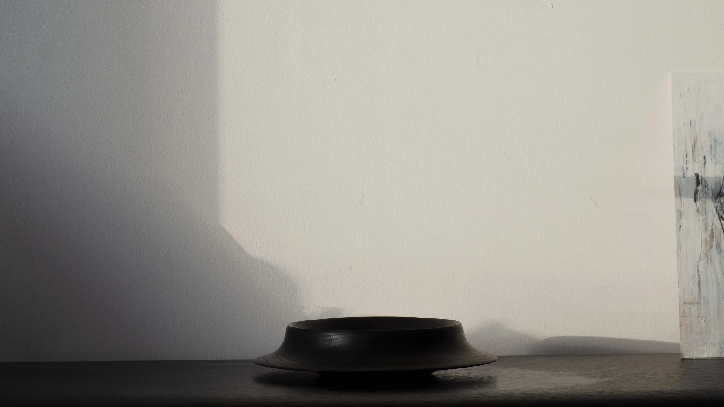 A minimalist scene featuring a dark pot on a flat surface against a plain wall with a small portion of artwork visible on the right side.