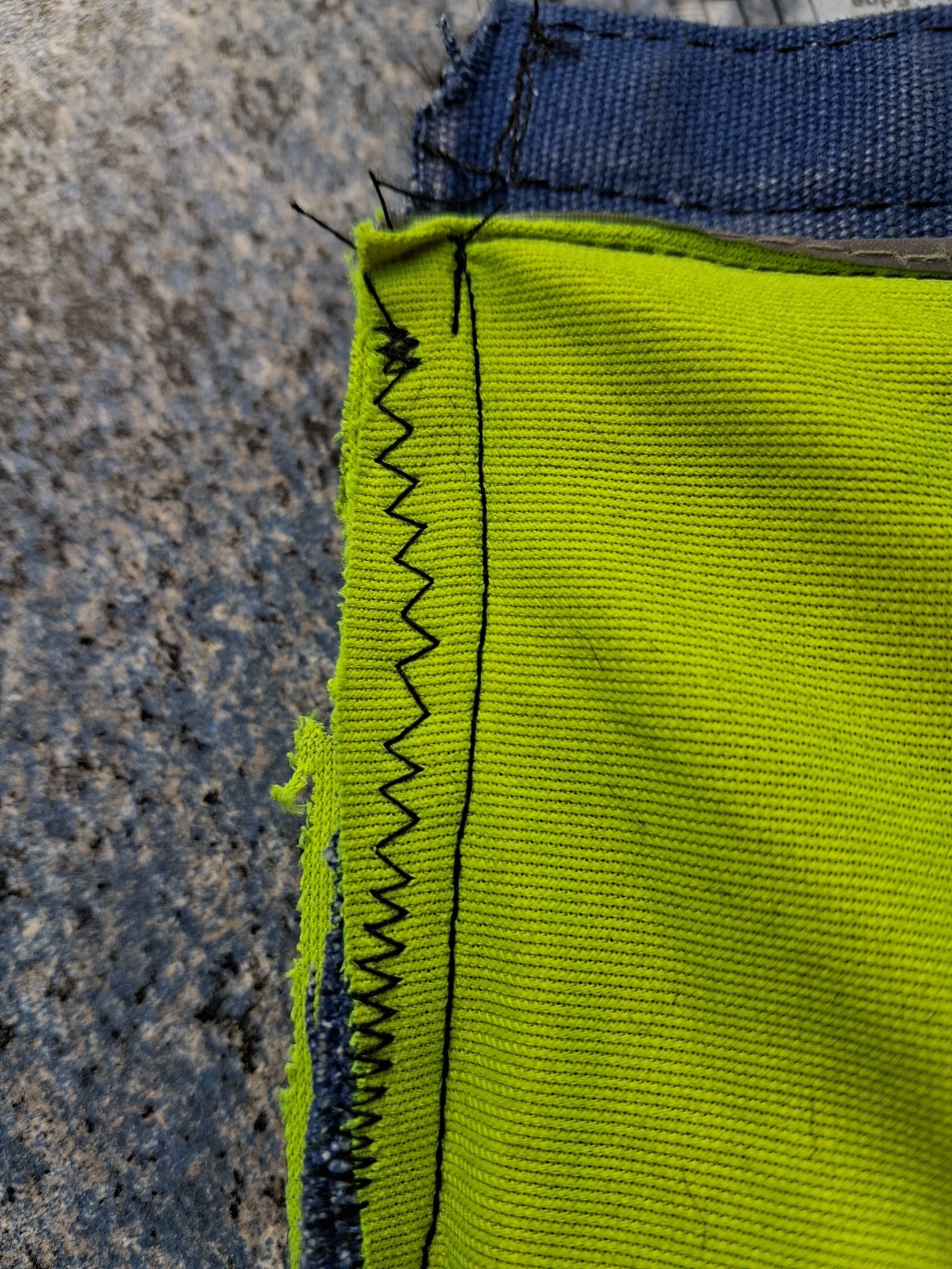 Close-up of a neon yellow work glove with black trim and black zigzag stitching on the cuff, worn on a person's hand.