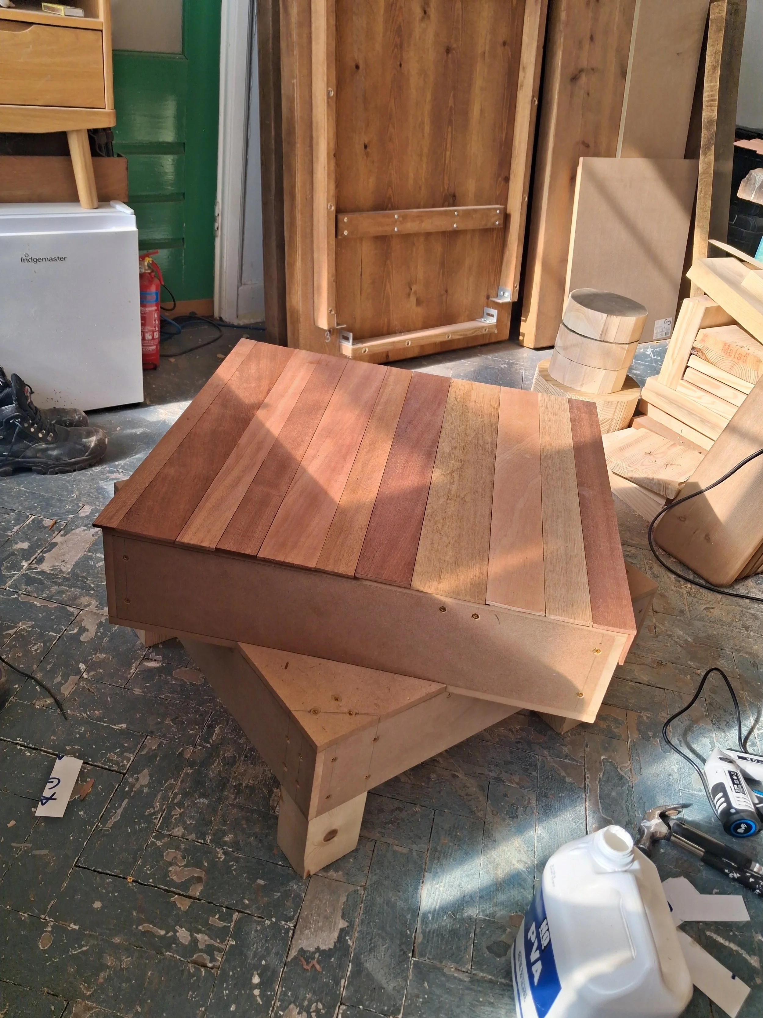 A wooden bench with a curved, paneled top sits on a workshop floor. Nearby, various tools and supplies, including a bottle of glue and a cordless drill, are visible. In the background, there are additional pieces of wood and a large, green door.