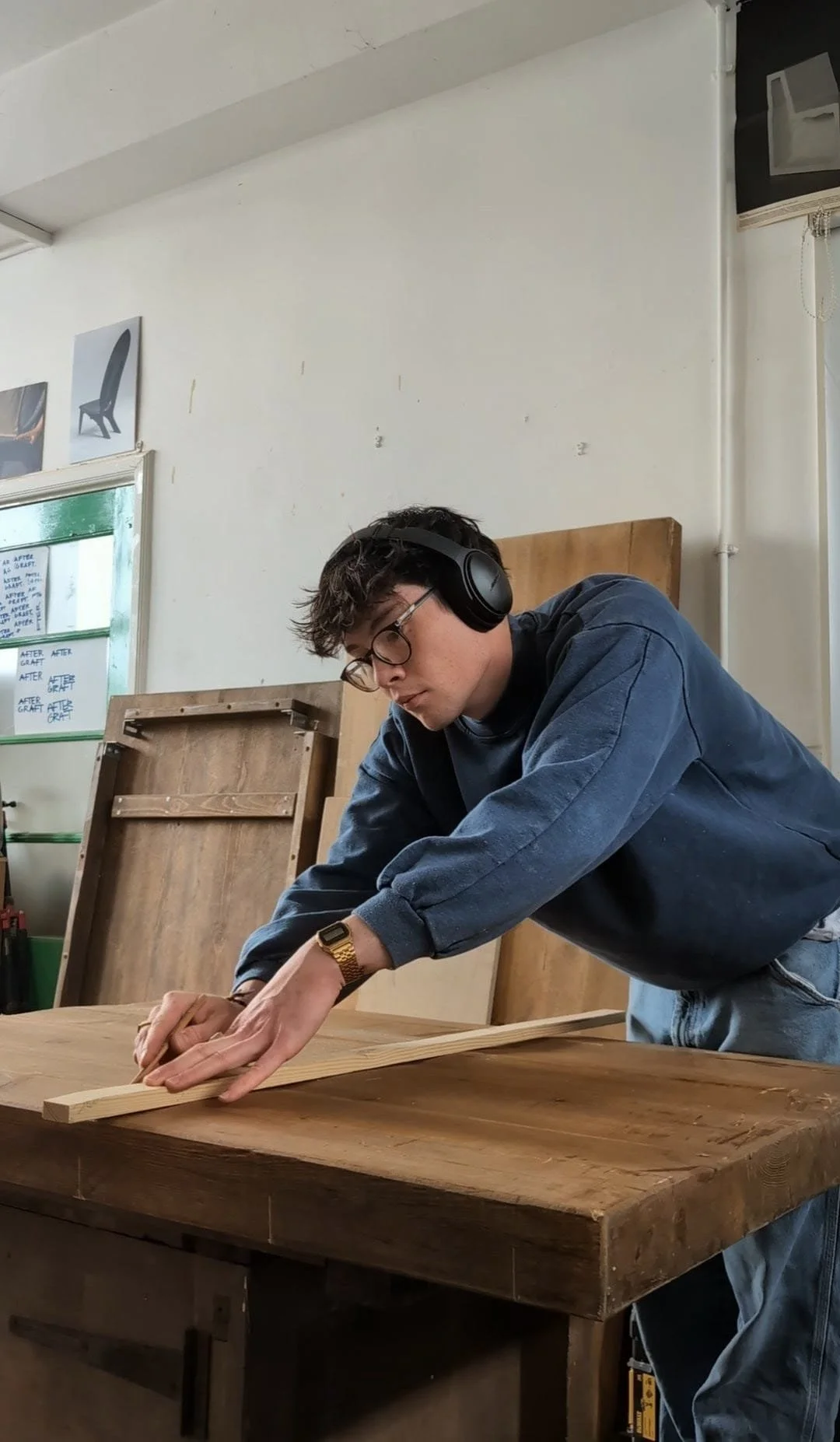 Young man cutting wood in a woodworking shop, wearing headphones and glasses, with wooden furniture and tools in the background.