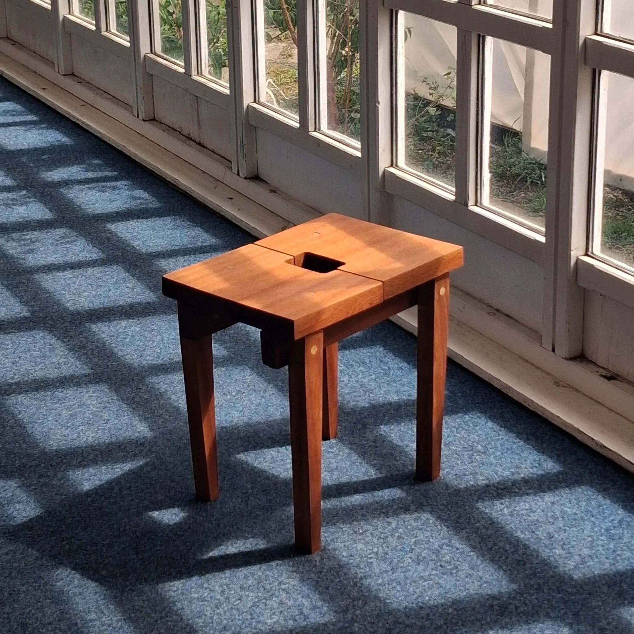 A small wooden table with a hole in the center is placed on a blue carpet near a large window with multiple panes. Sunlight creates shadows on the carpet inside a room.
