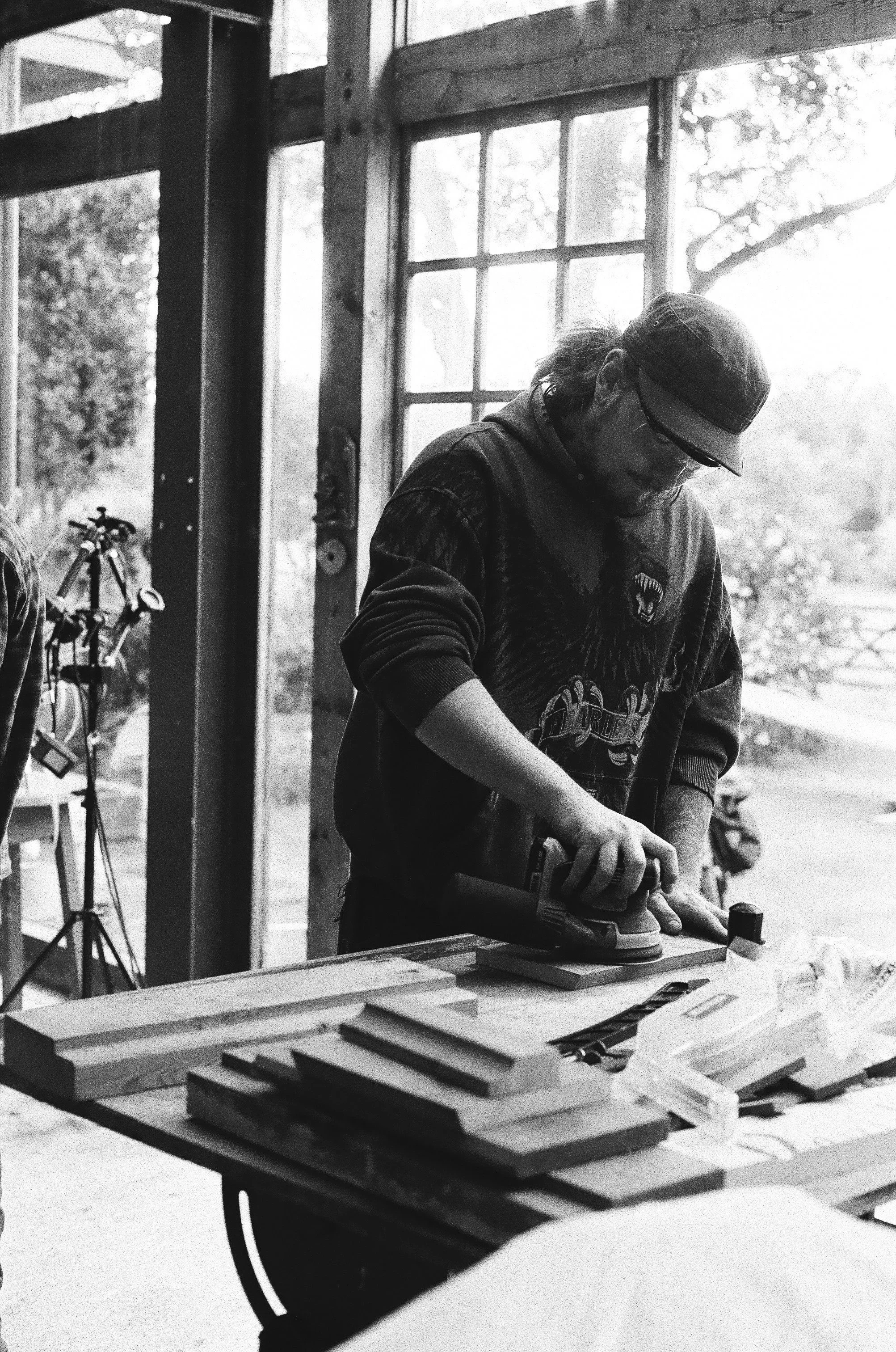 A person wearing glasses, a cap, and a hoodie working with a power sander on a piece of wood inside a rustic room with large windows and trees visible outside. A skateboard and stacks of wooden pieces are on the table in the foreground.