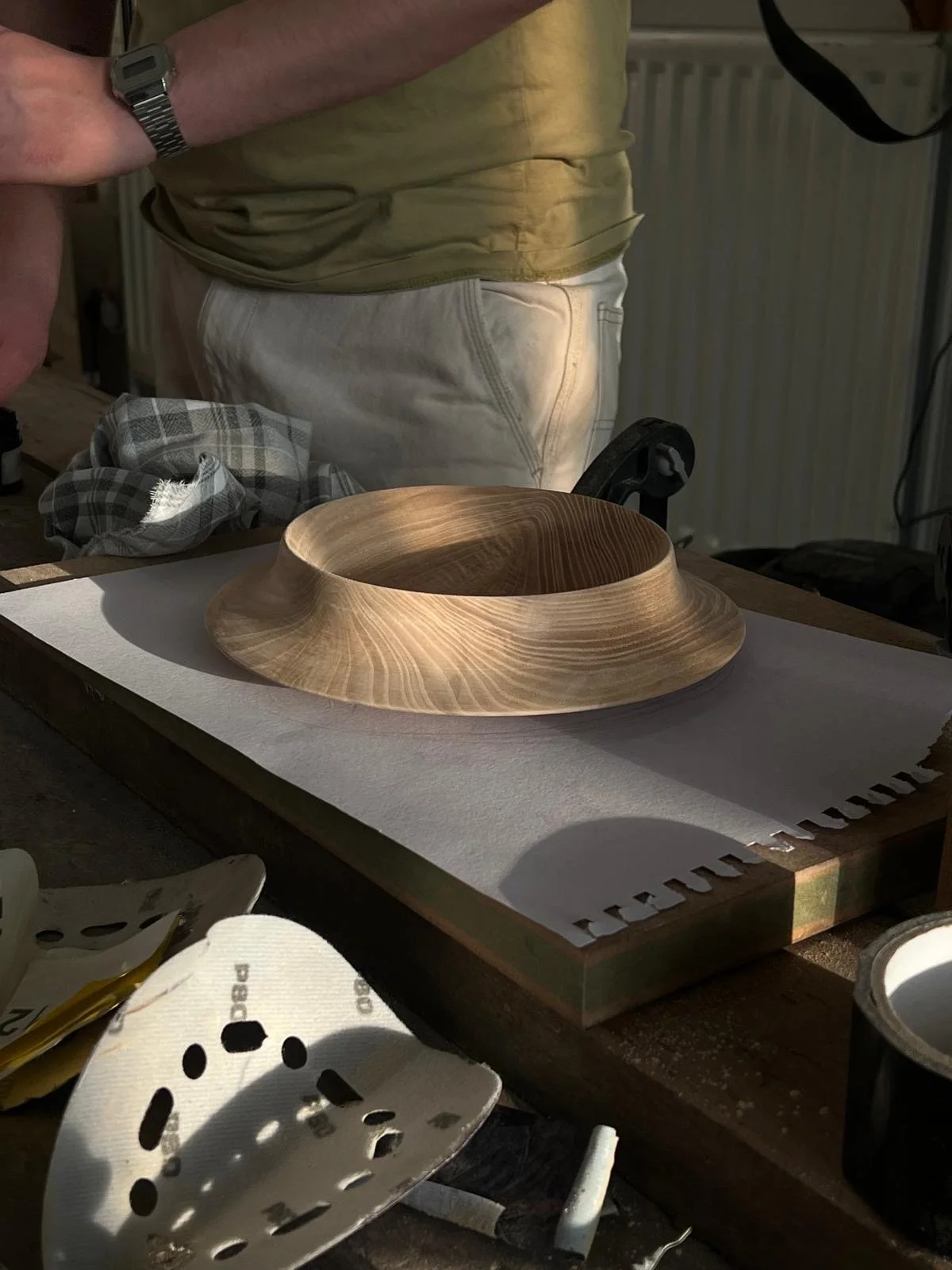 A wooden bowl with a smooth, curved design is placed on a work table in a woodworking shop. A person wearing a yellow shirt and white apron is working nearby, with woodworking tools and a cloth visible on the table.