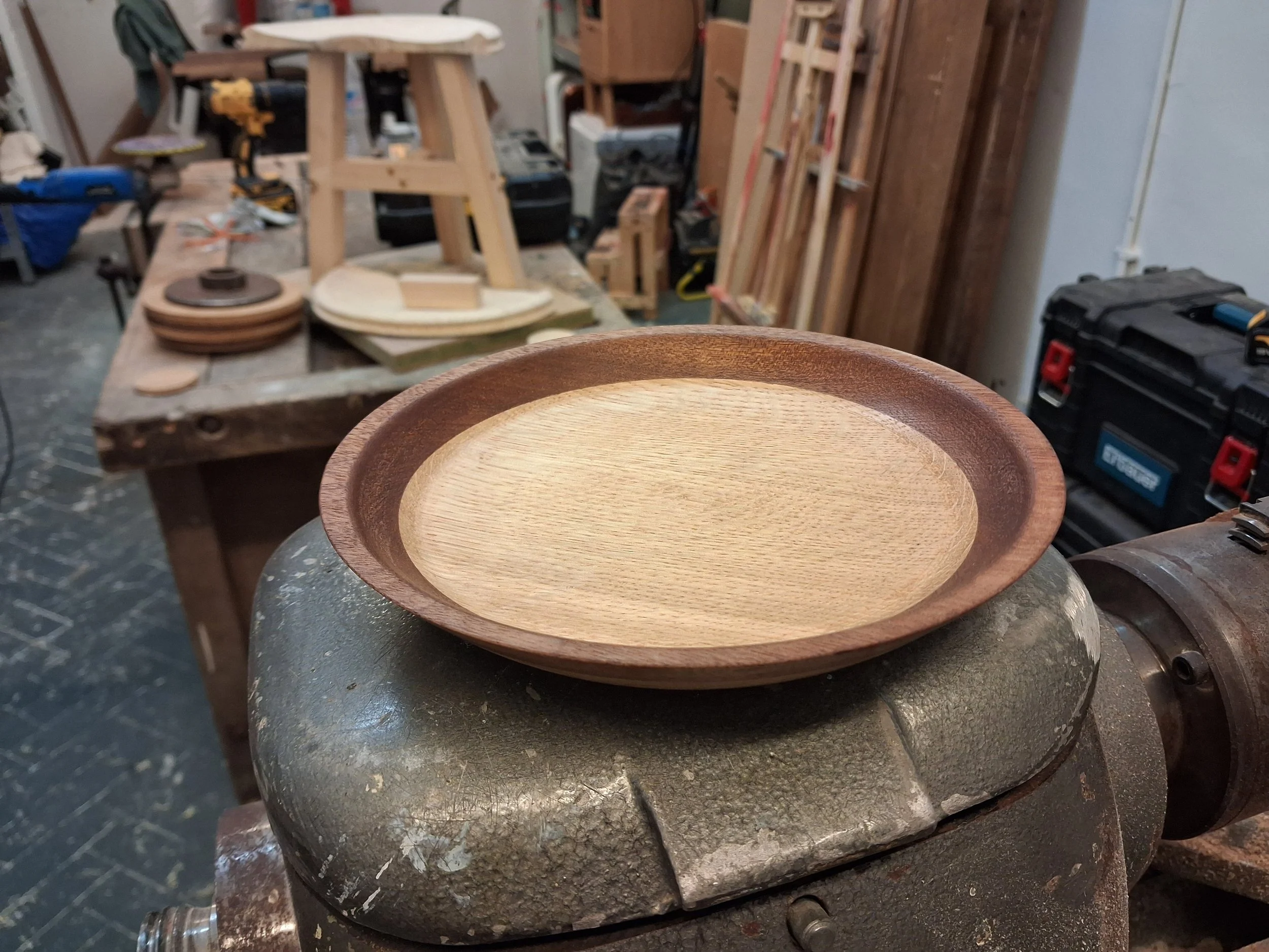 A woodworking workshop with a bench, tools, and wood pieces. In the foreground, a half-finished wooden bowl is held on a workbench. The background shows woodworking supplies and materials.