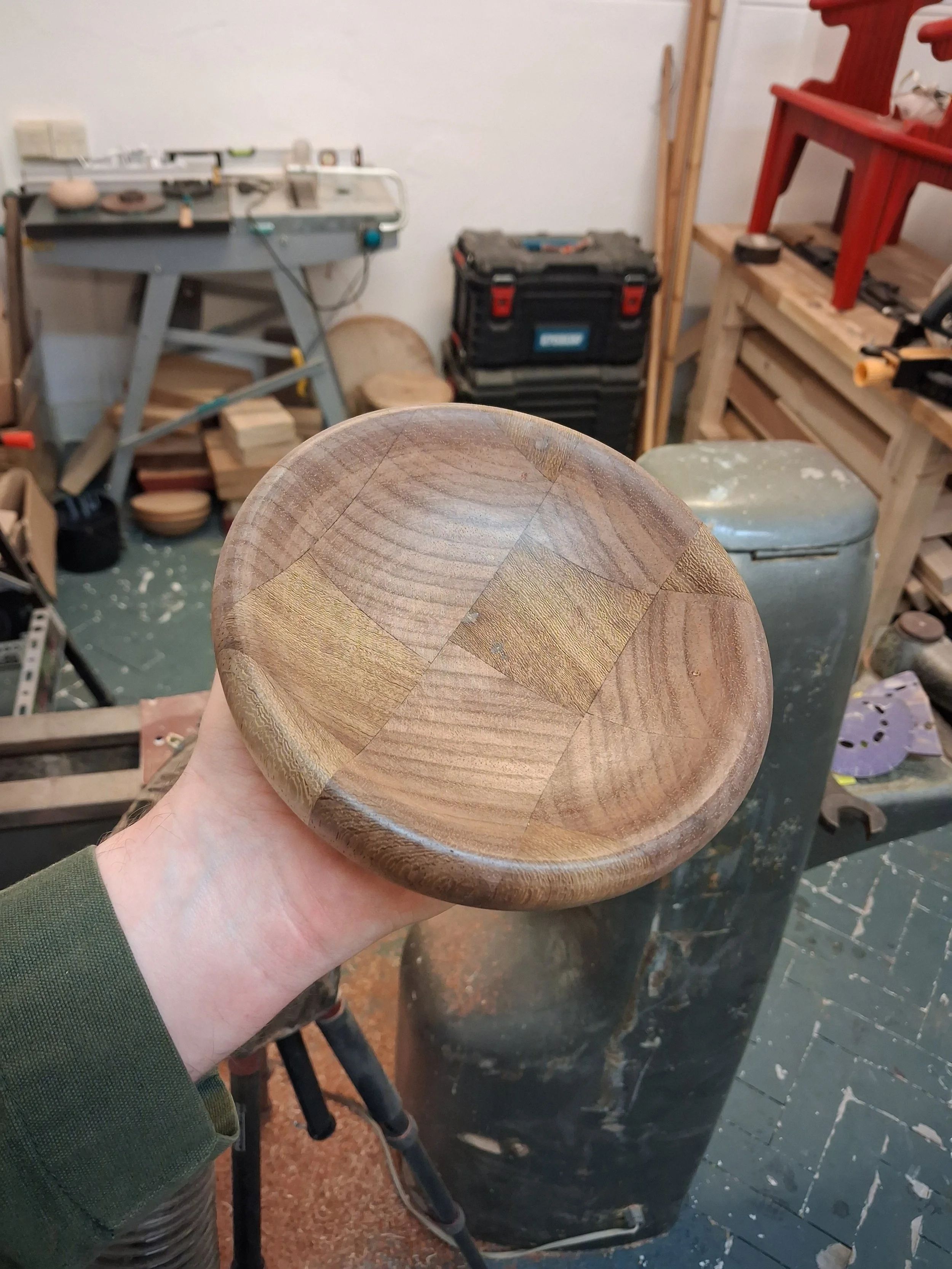 A wooden bowl being held by a person in a woodworking shop, with workbenches, tools, and stacks of wood visible in the background.