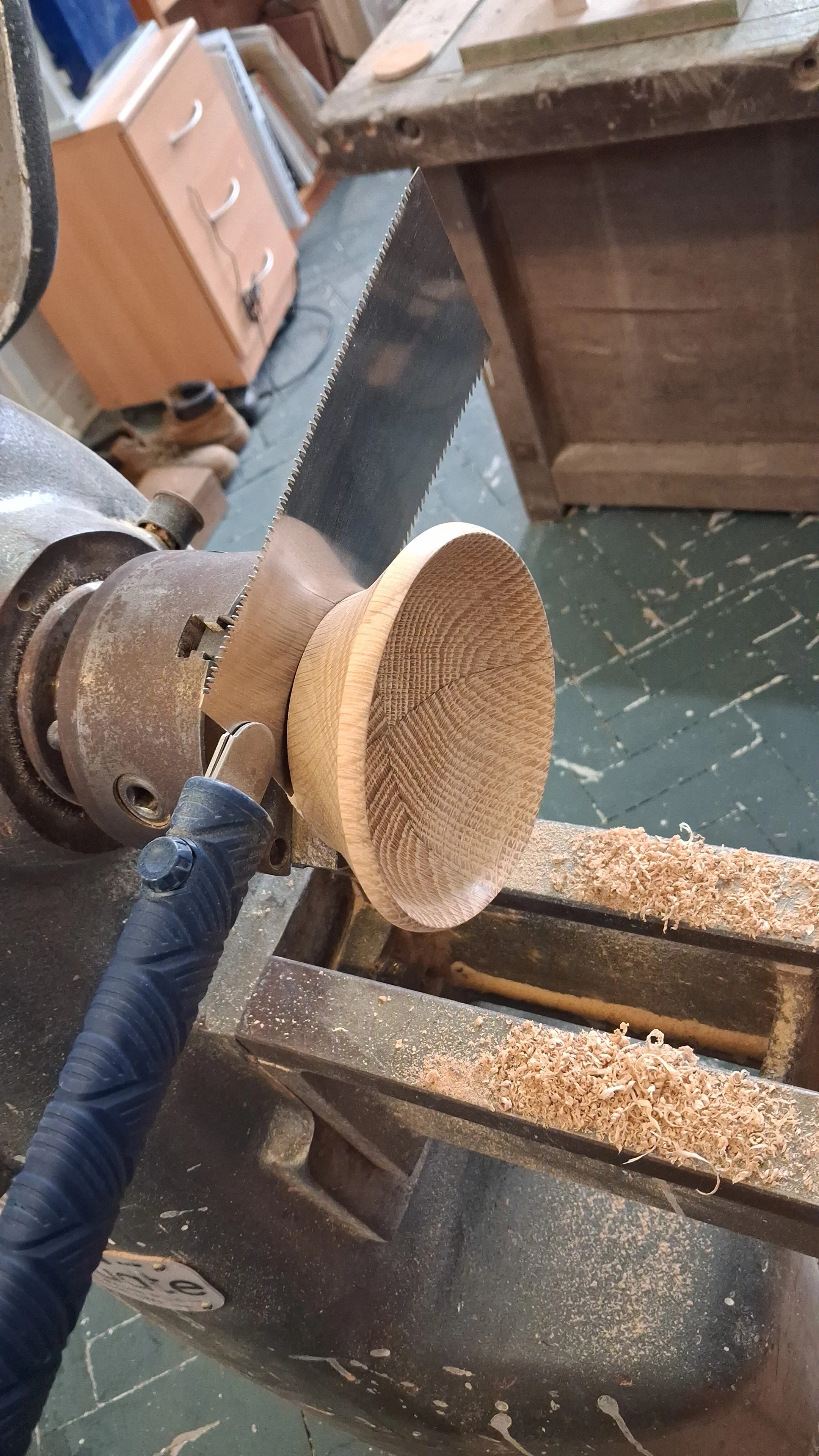 A woodworking shop showing a close-up of a wood lathe with a piece of turned wood, a bandsaw blade, and sawdust on the surface.