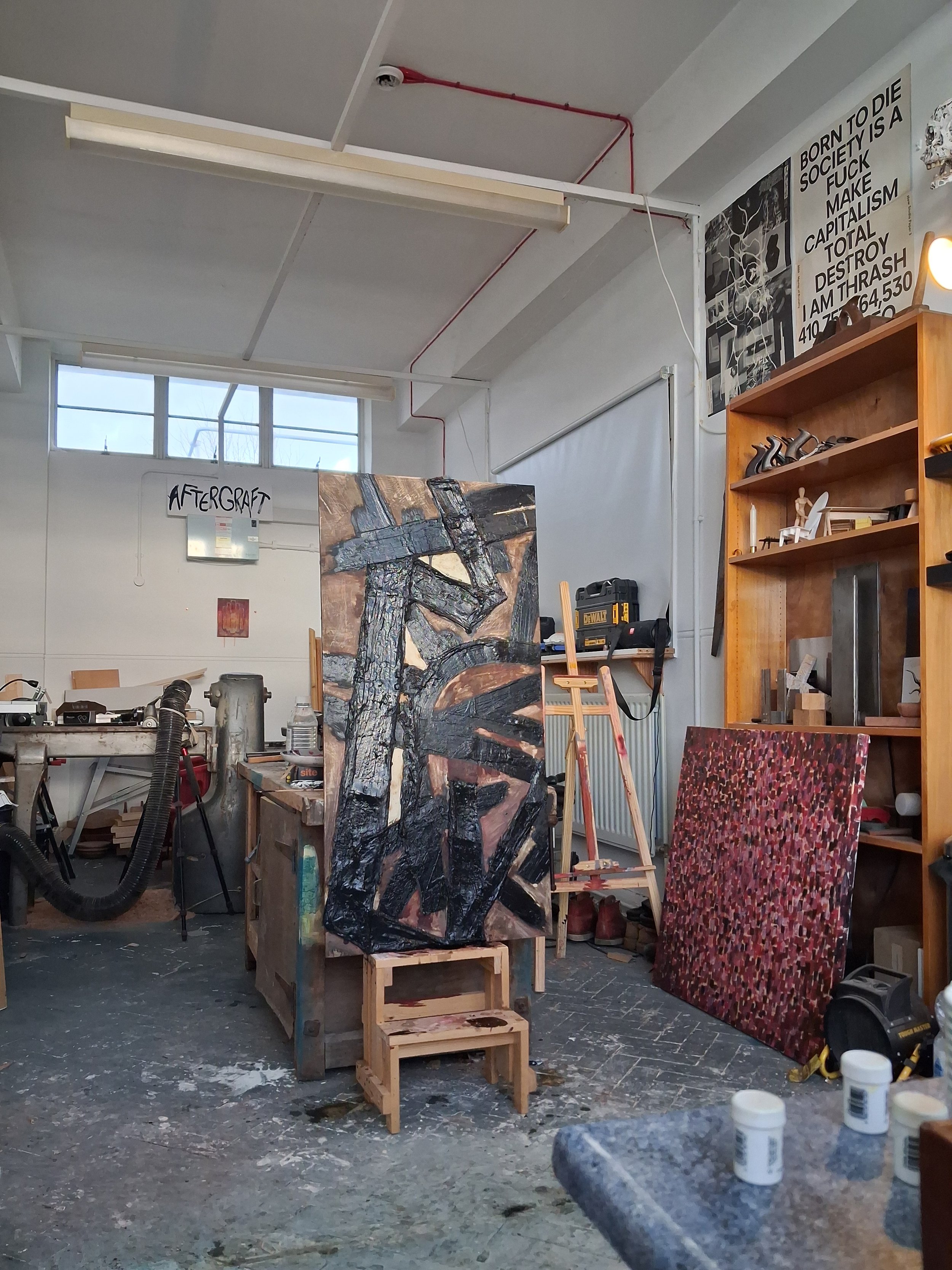 An art studio with paintings, art supplies, and furniture including a colorful abstract piece, a wooden shelf with sculptures, paint bottles, and a workspace with tools and a vacuum cleaner.