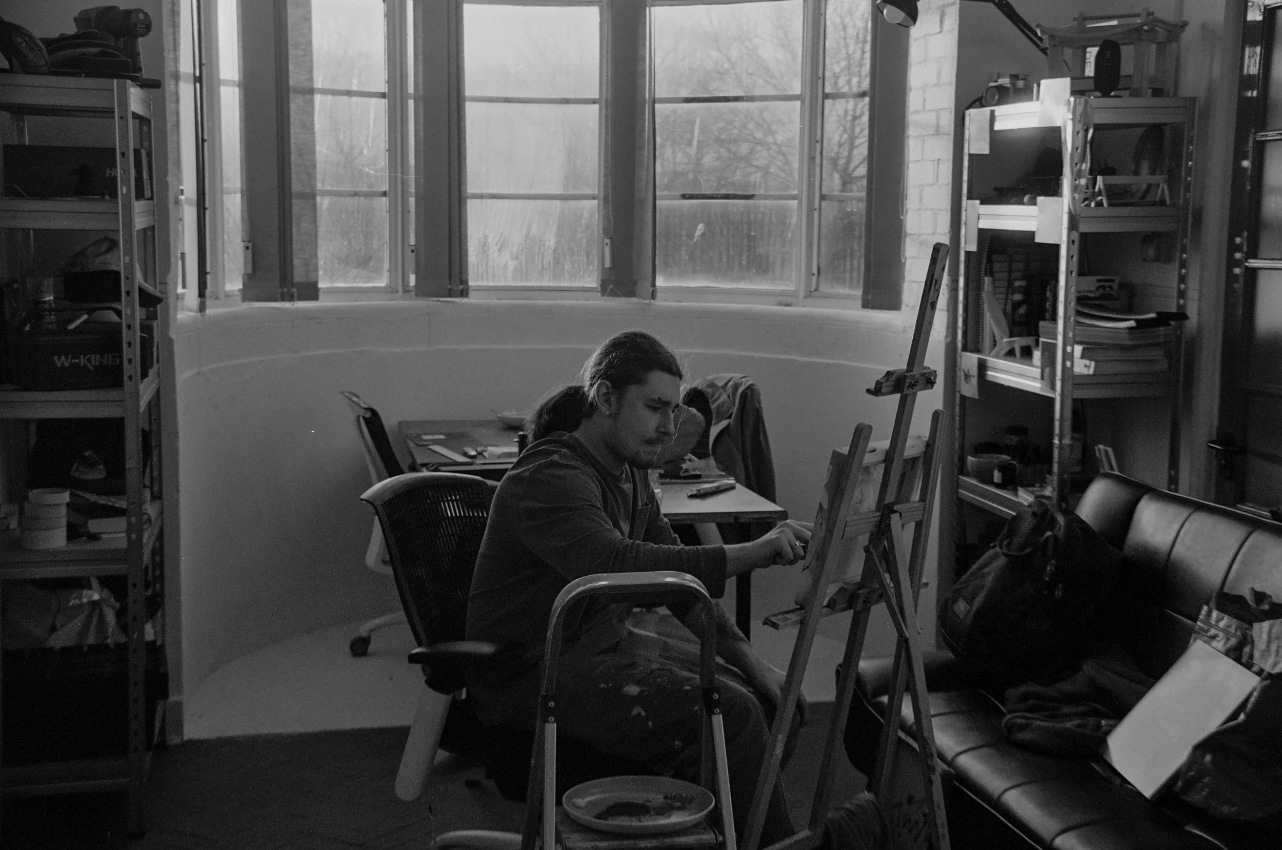 A man working on an easel in a room with large window and metal shelves.