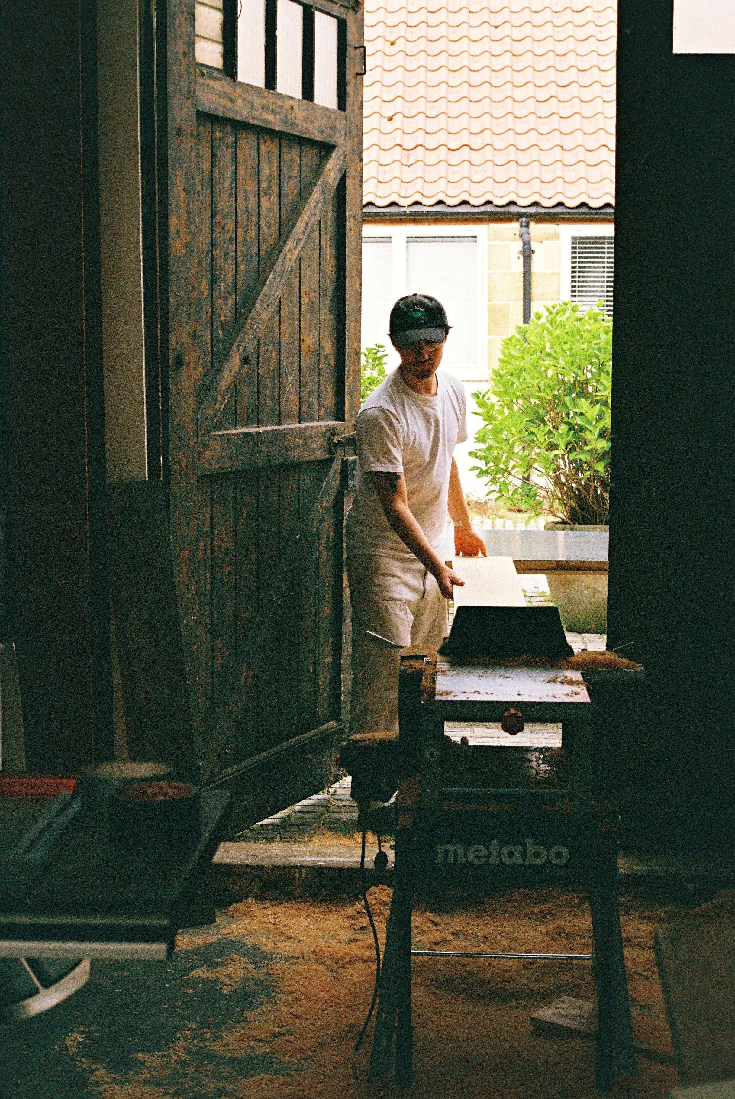 A man wearing a white t-shirt, white shorts, and a black cap is standing outdoors in front of a wooden barn door that is open to reveal a garden with green bushes and a house. A metal workbench with a saw and other tools is in the foreground, and the ground is covered with sawdust.