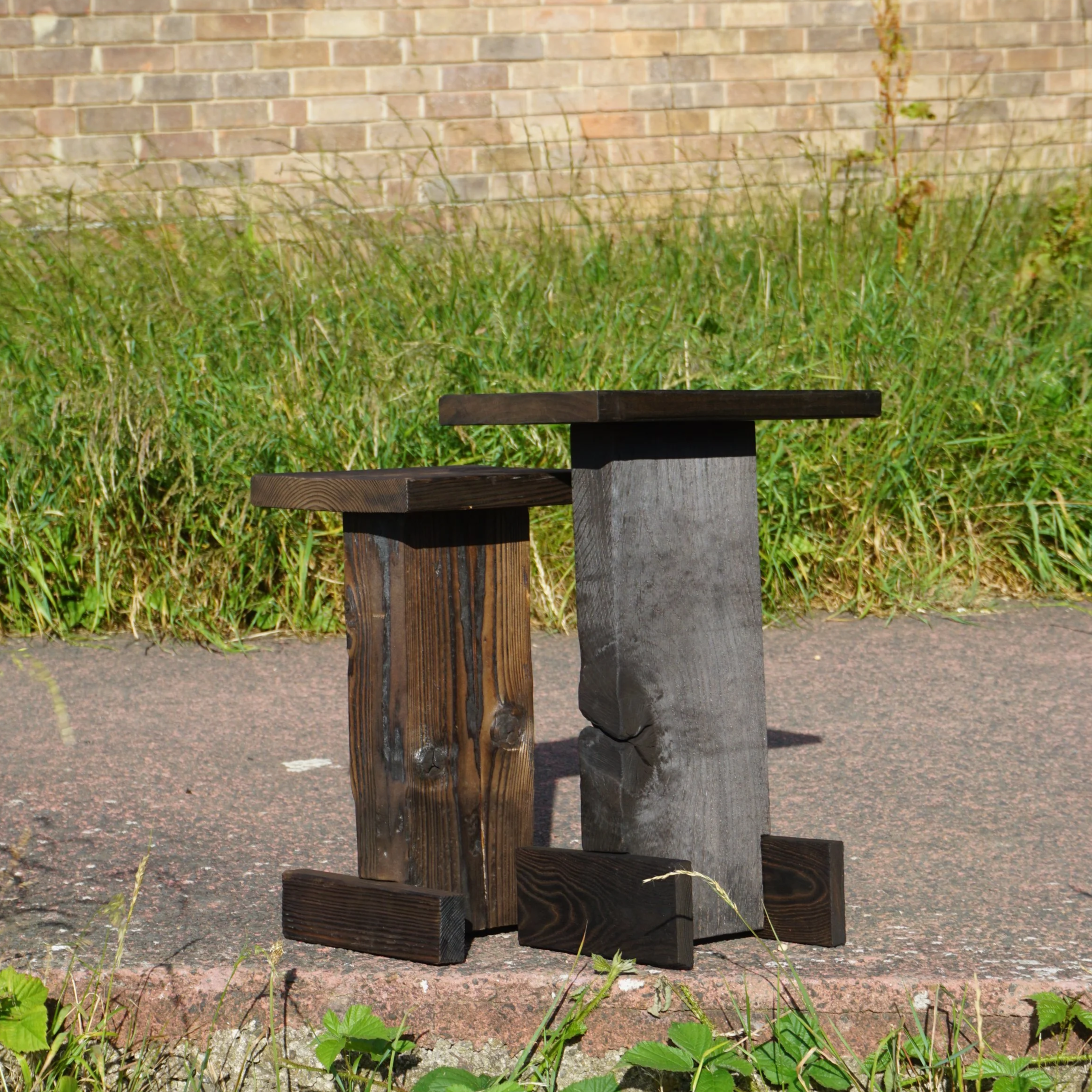 Several dark-colored wooden blocks arranged to form a small sculpture or structure, placed on the ground with surrounding green plants and a brick wall in the background.