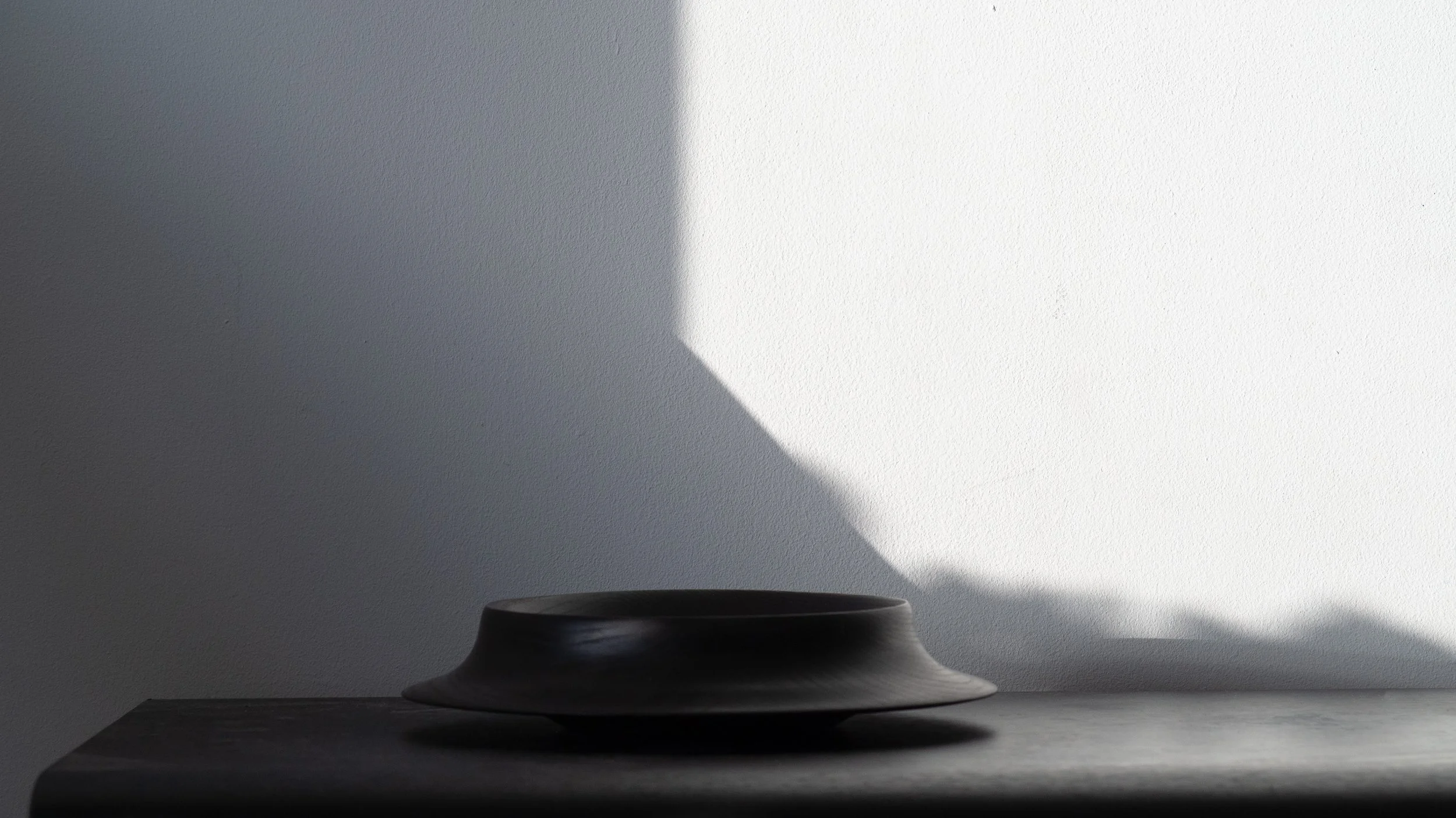 A ceramic bowl on a dark surface with sunlight casting a shadow on a textured wall behind it.
