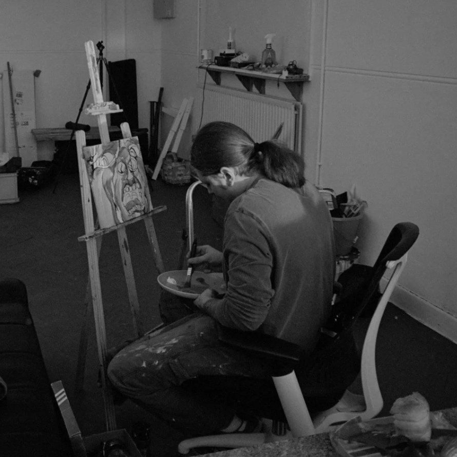 A woman with a ponytail, wearing a jacket, sits on a chair in an art studio painting a canvas on an easel. The canvas depicts an abstract face with exaggerated features. The room has art supplies, shelves, and a radiator along the wall.