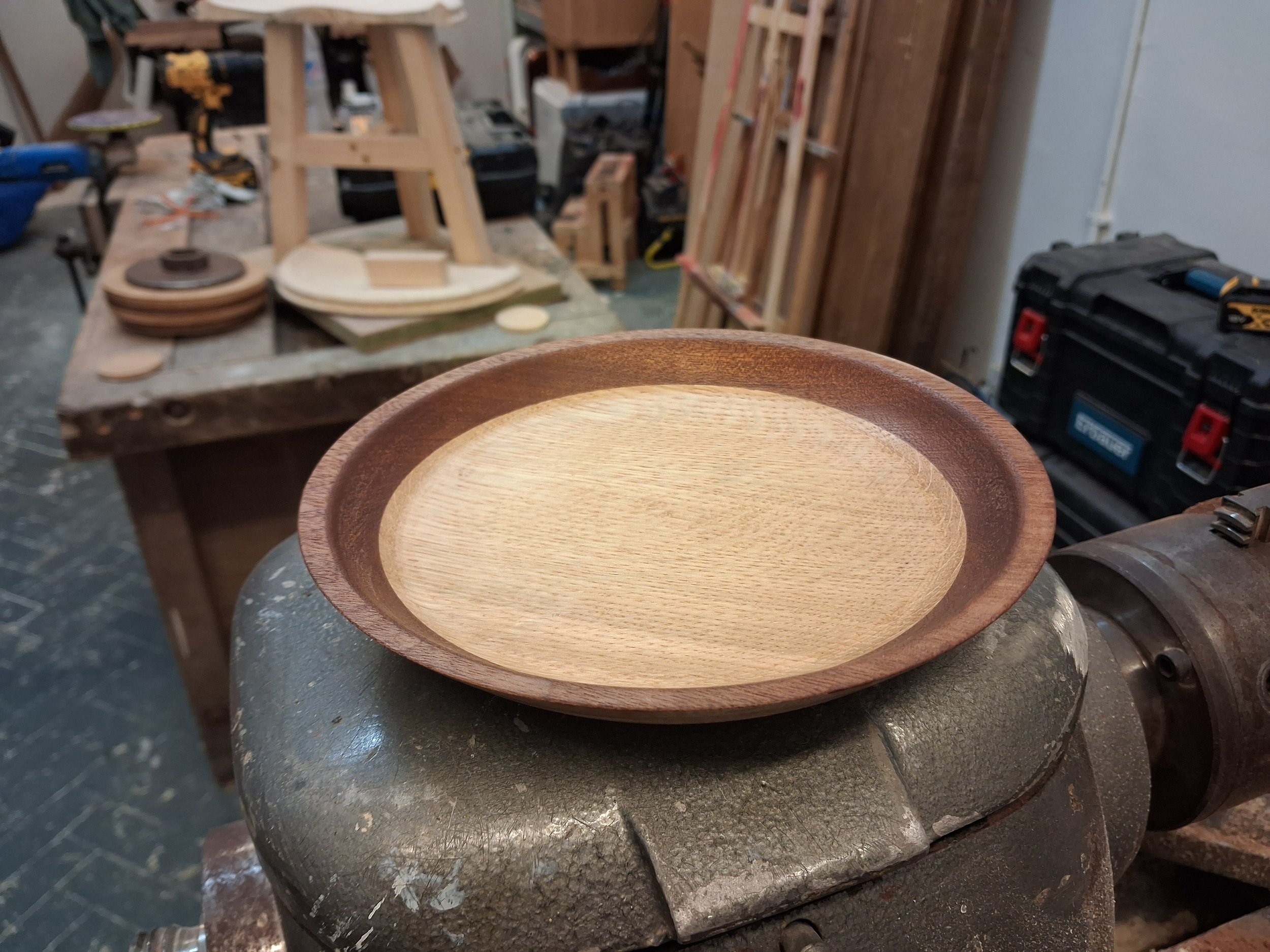 A wooden bowl with a light inner surface and darker exterior surface in a woodworking shop. The background shows woodworking tools, stacks of wood, and a workbench.