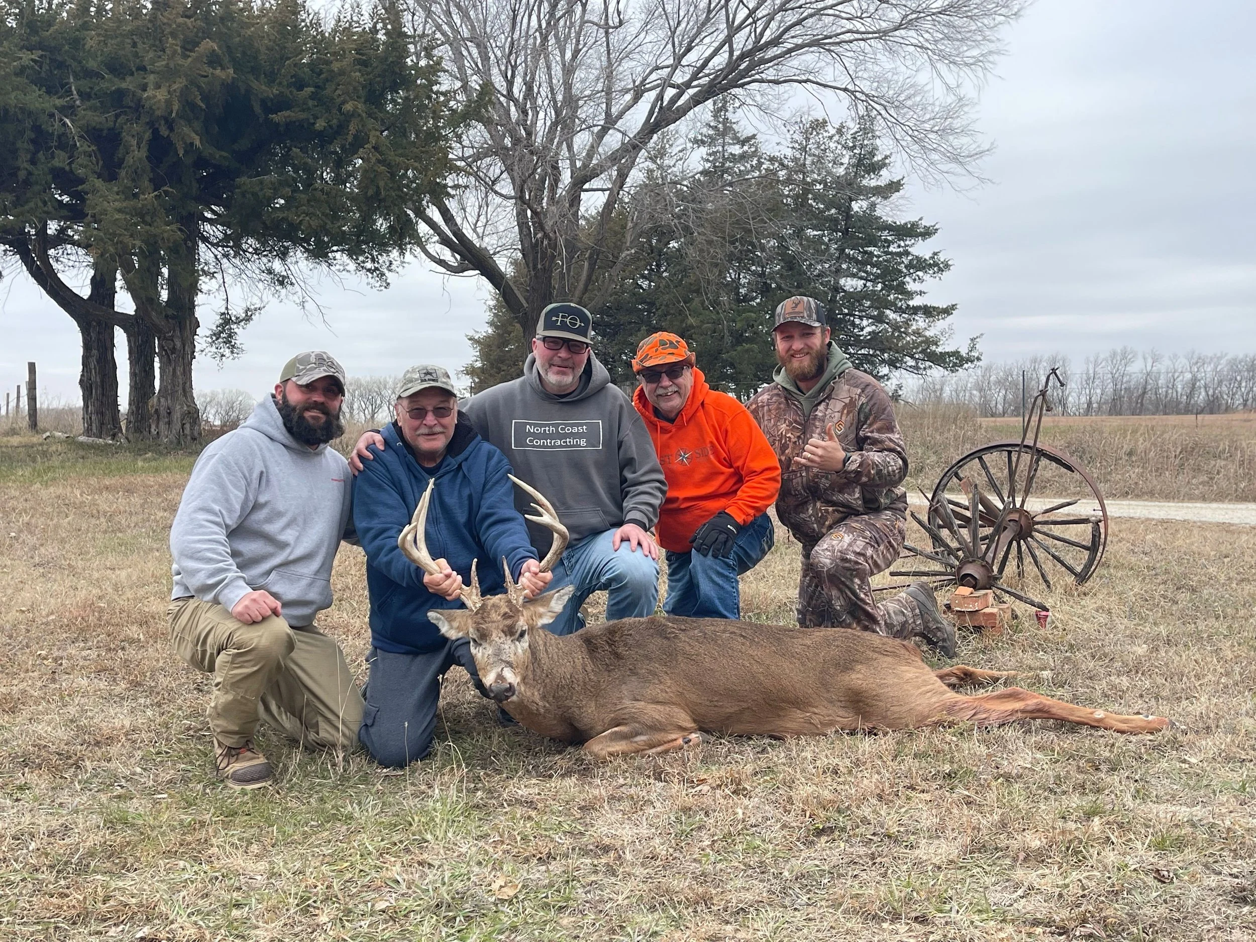 Five men kneeling and standing around a harvested deer with antlers, outdoors in a field with trees in the background, and an old wagon wheel on the ground.