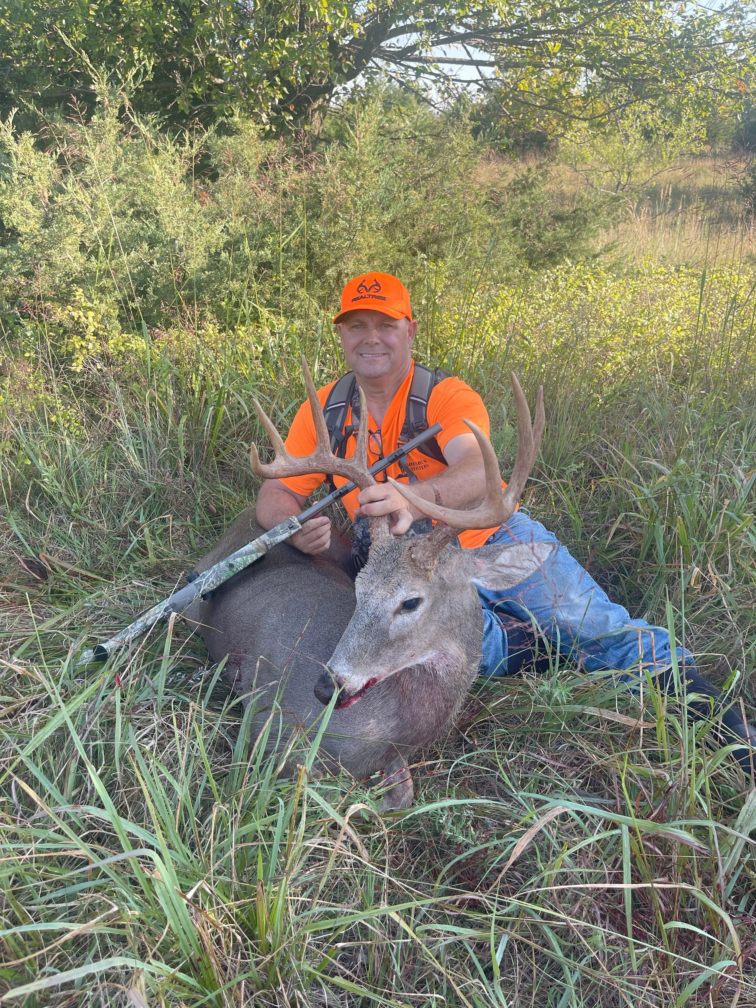 A man in orange hunting gear sitting with a large deer in tall grass during daytime.