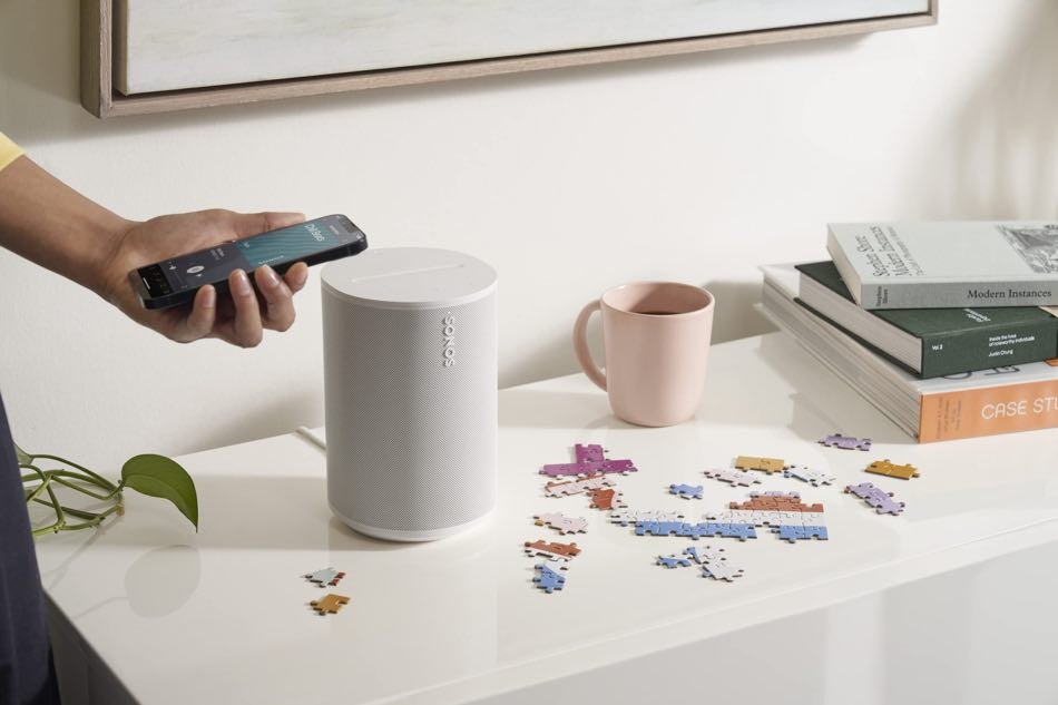 Una mesa blanca con una mano que sostiene un teléfono móvil, una bocina blanca, una taza rosa, algunos rompecabezas en piezas y varios libros apilados.