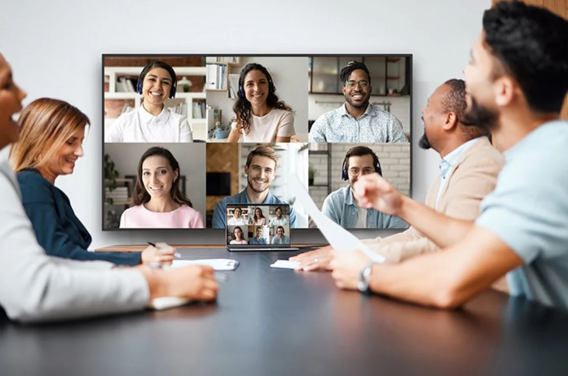 Reunión de varias personas en una videoconferencia en una pantalla grande, con algunas personas tomando notas.