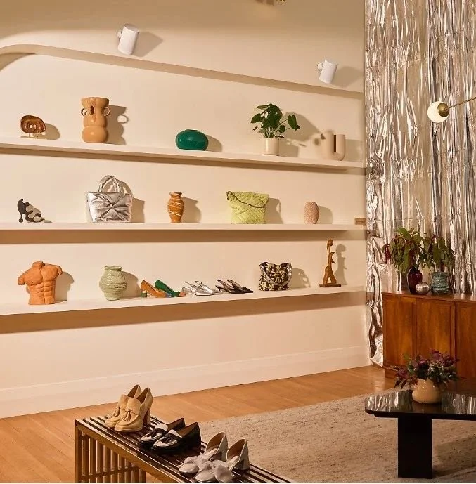 Estanterías blancas con objetos decorativos en la pared, incluyendo jarrones, plantas, bolsas y esculturas, en un salón con piso de madera y cortinas plateadas.