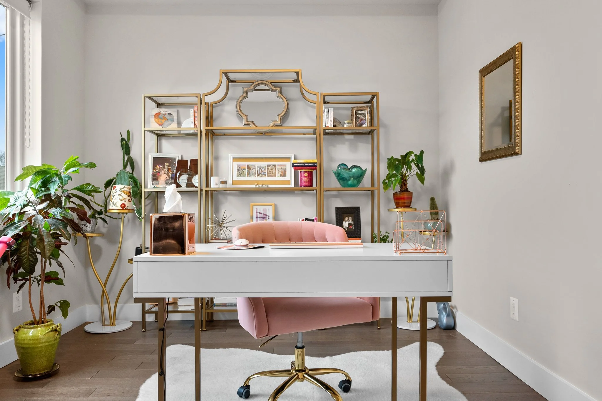 A home office with a white desk, pink swivel chair, gold shelving with decor, framed pictures, and plants, including a large potted plant on the left and a small plant on the right. A gold-framed mirror hangs on the wall to the right.