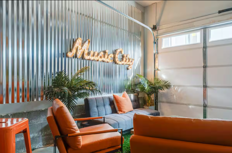 A garage decorated with a grey couch, orange chairs, and pillows with a lit sign saying," Music City"
