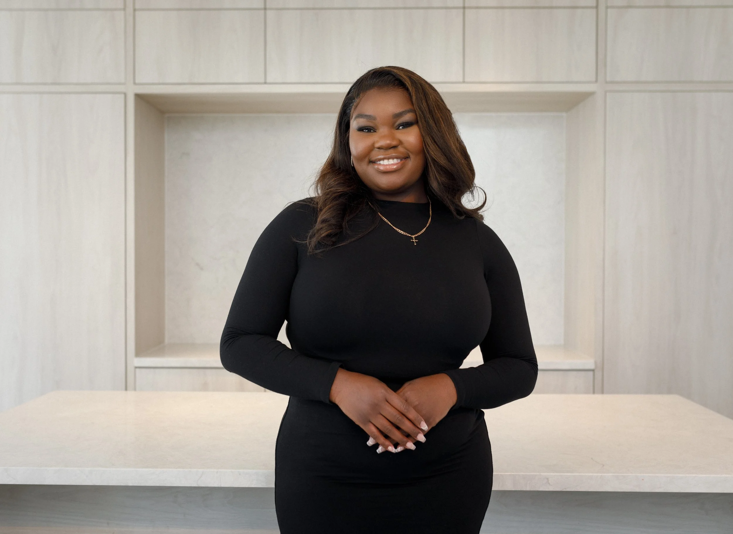 A woman in a black long-sleeve dress standing indoors with a neutral background and white countertop.
