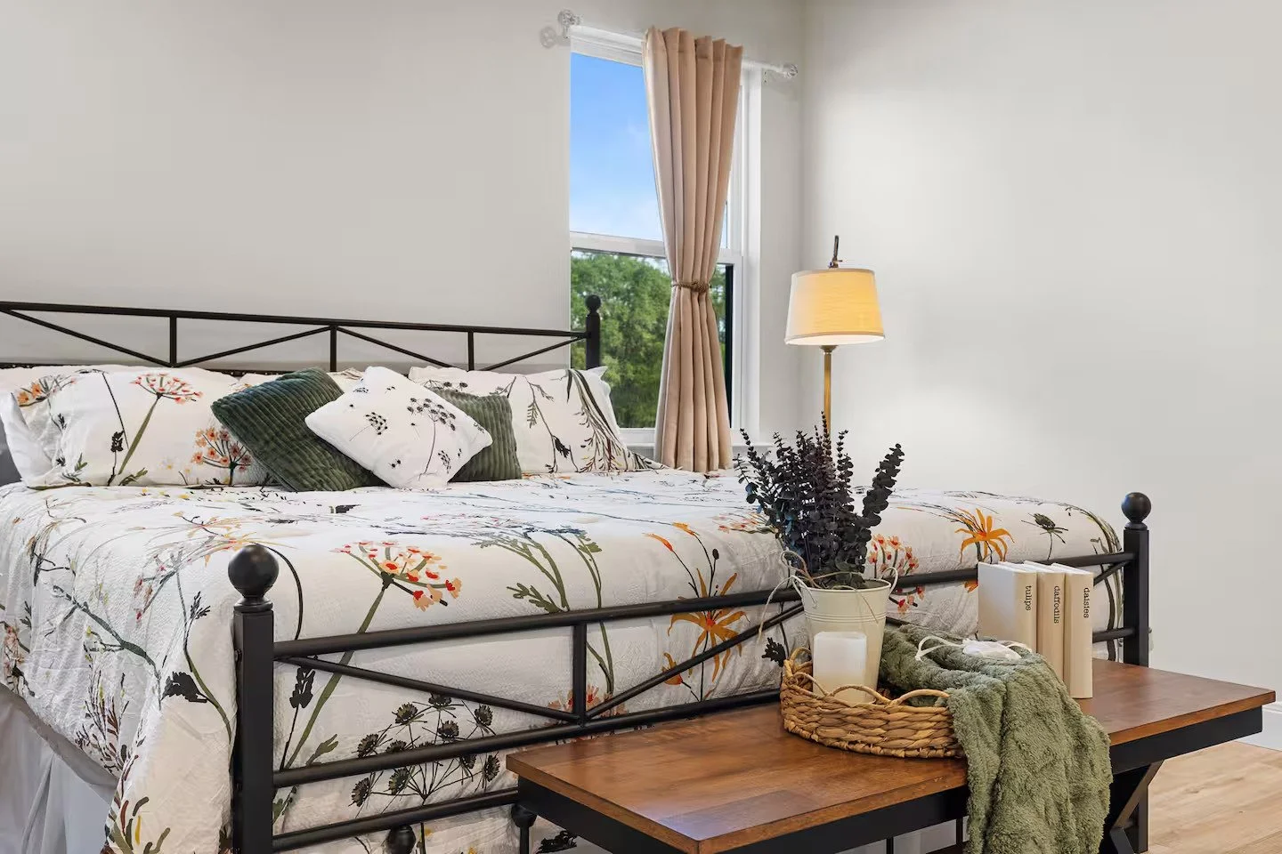 A bedroom with a large bed covered in white floral bedding and pillows, a wooden side table with a plant, candles, and books, and a window with beige curtains allowing natural light.