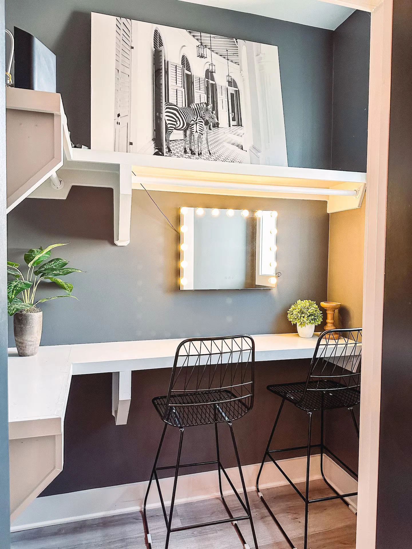 A view of built-in wall shelving used as a desk with two bar stools in front and a small mirror to do your makeup.