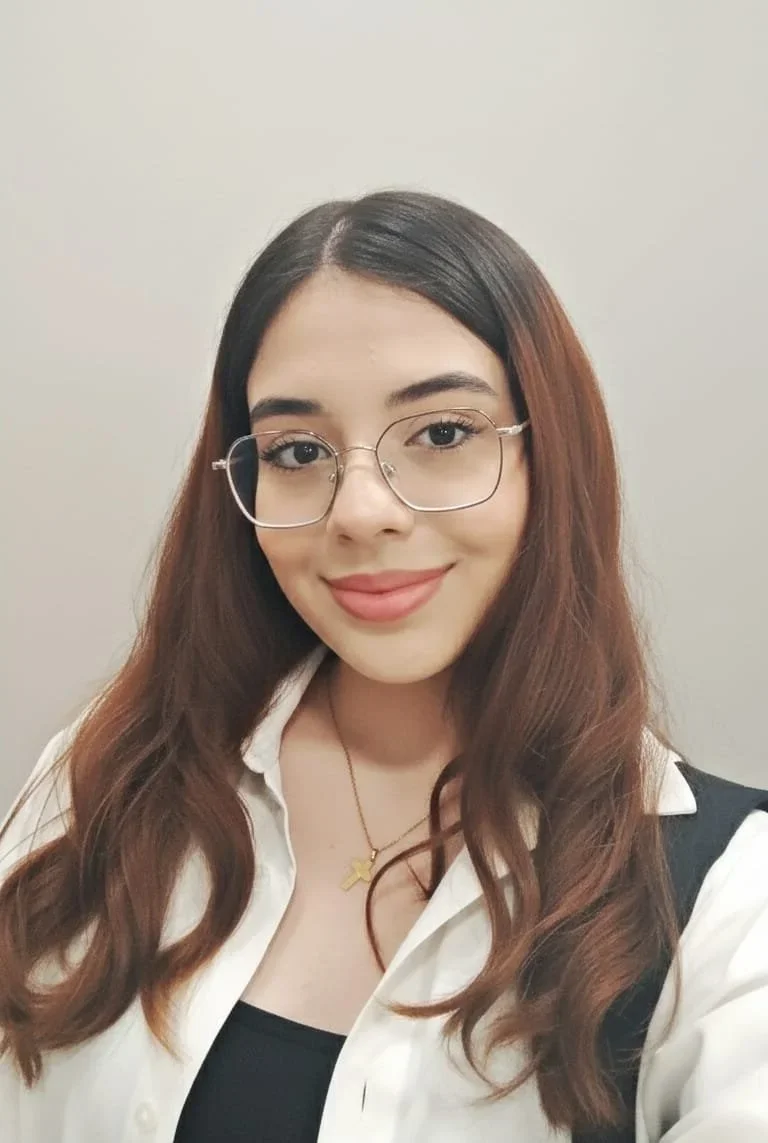 A young woman with long wavy brown hair, wearing glasses, a white shirt, a black top, and a gold cross necklace, smiling at the camera.