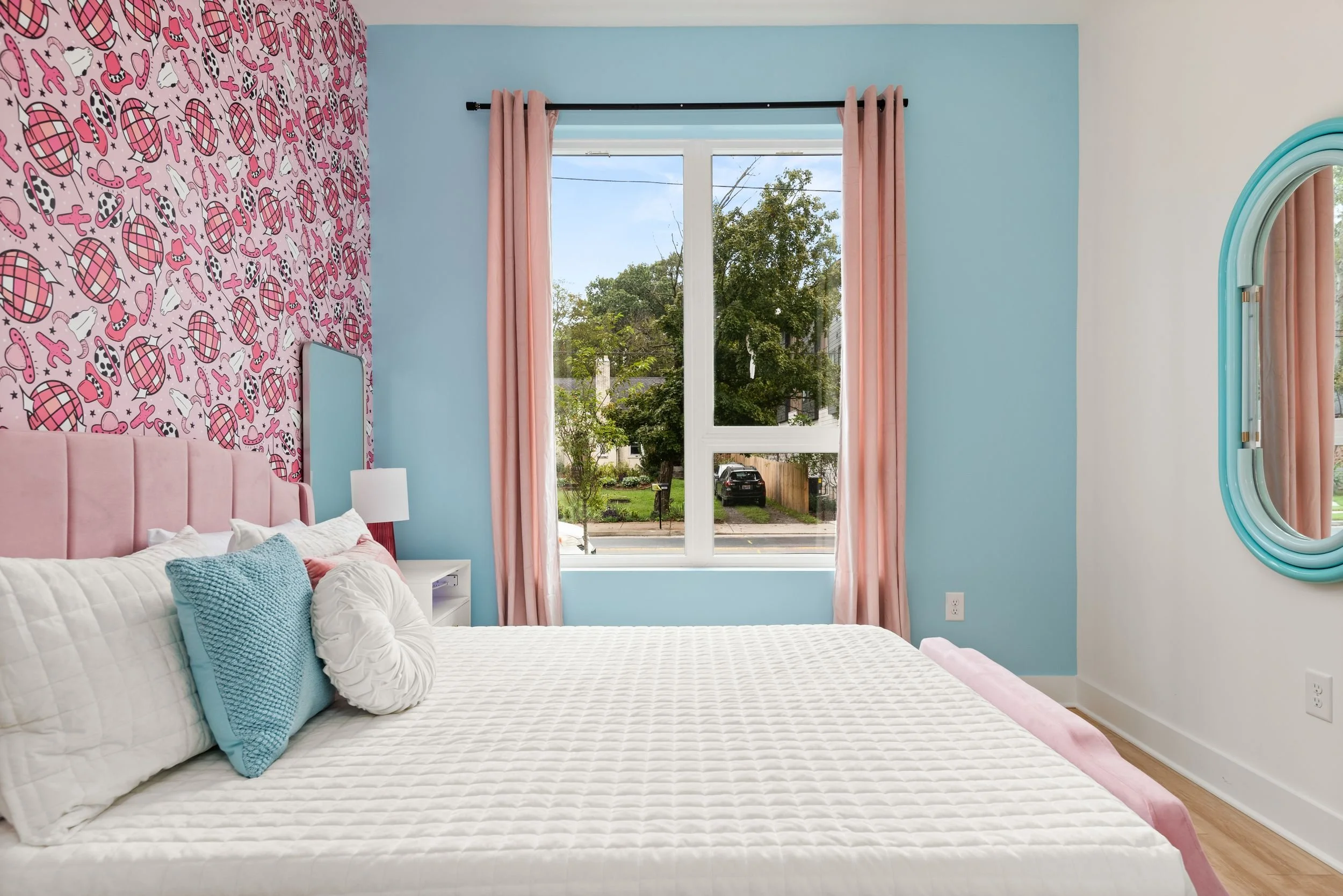 An aqua blue bedroom with a pink bedframe bed with white, pink, and blue bedding, pink curtains, and one wall with cowboy disco wallpaper.