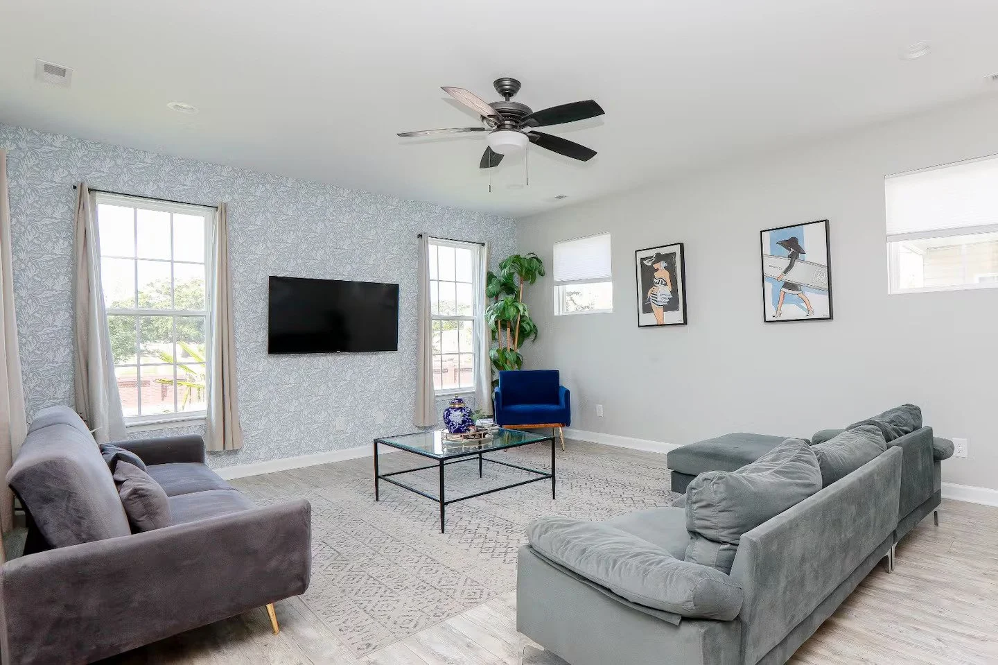 A living room view of two grey sofas and a velvet blue chair surrounding a black frame, glass coffee table on a grey patterned rug in front of a tv.