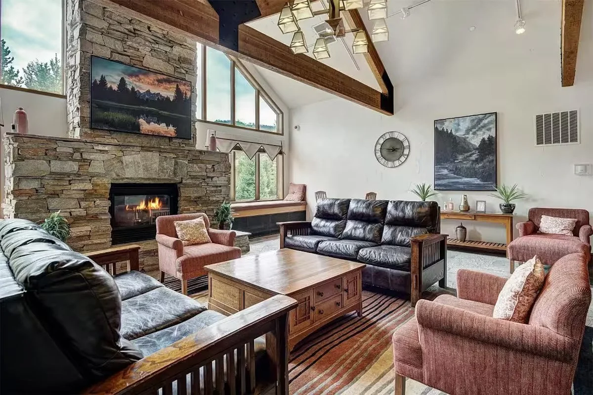 A living room with pink chairs, two brown sofas, a large square coffee table, tall brick fireplace, and a large wooden beam running across the vaulted ceiling.