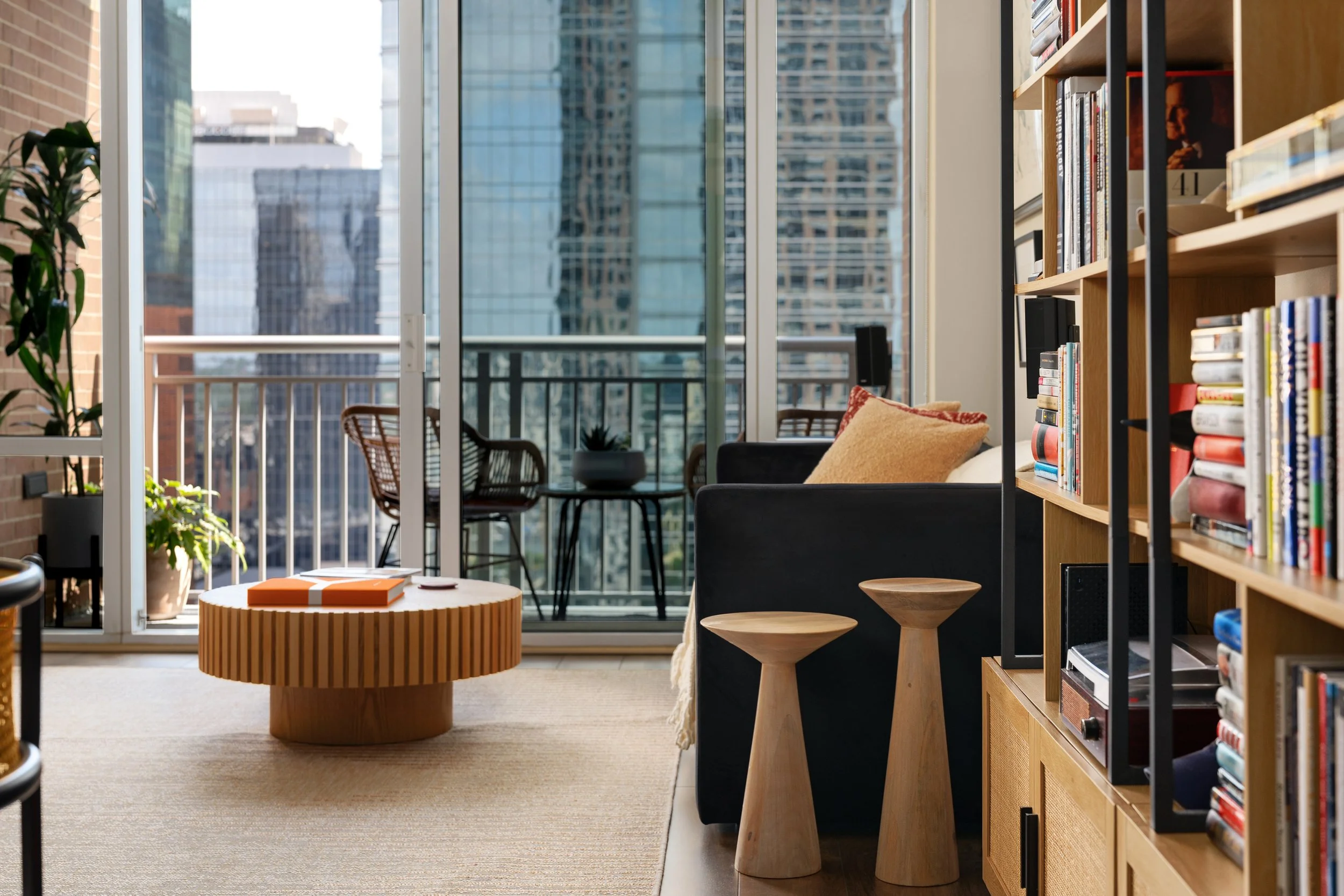 A city loft living room with a black sofa, round end tables and coffee table, and a balcony view of city buildings.
