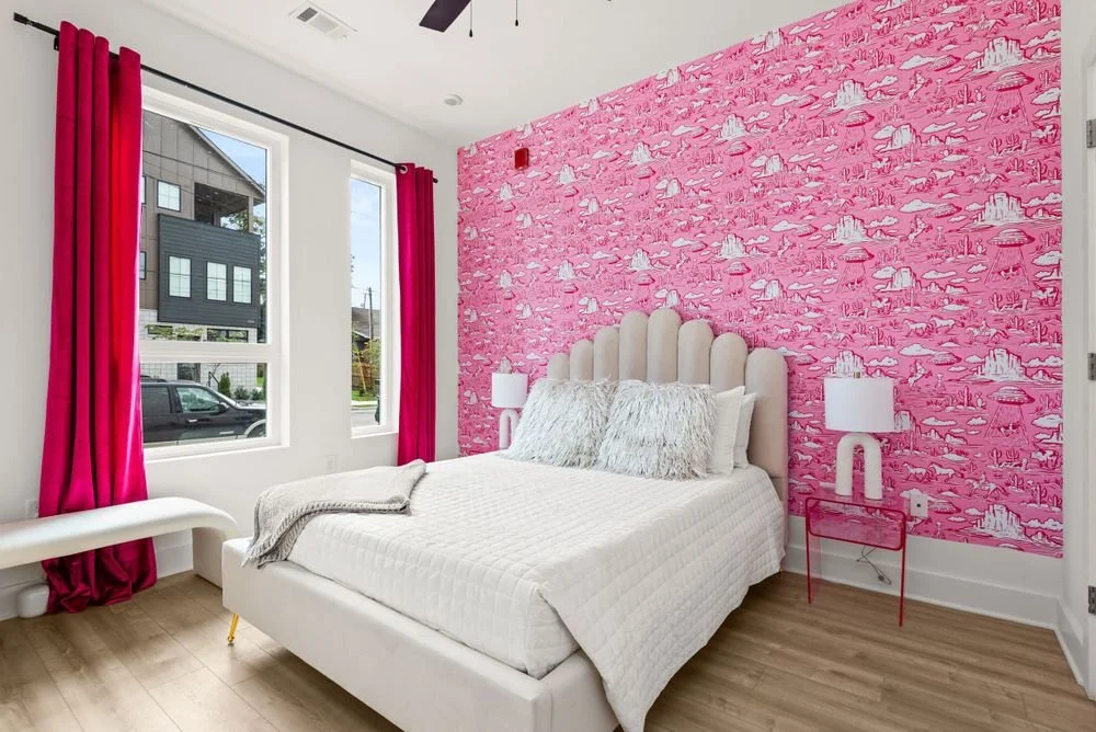 A bed with soft white and cream bedding and bed frame with two side tables, hot pink curtains, and is against a pink wallpaper wall.
