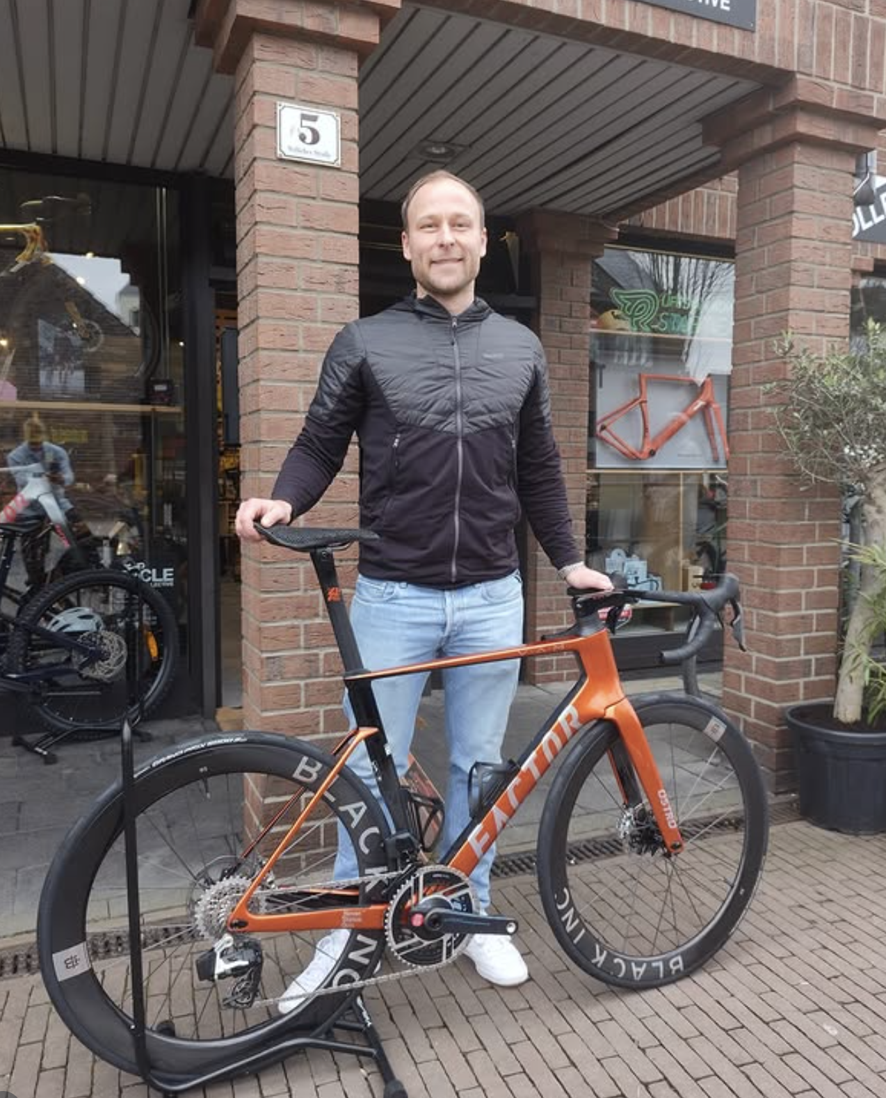 Ein Mann steht vor einem Fahrradgeschäft mit einem orangefarbenen Rennrad und lächelt. Das Geschäft befindet sich in einem rotem Backsteingebäude. Es gibt Fahrradzubehör im Schaufenster und ein Fahrradrahmen ist an der Wand hinter ihm montiert.