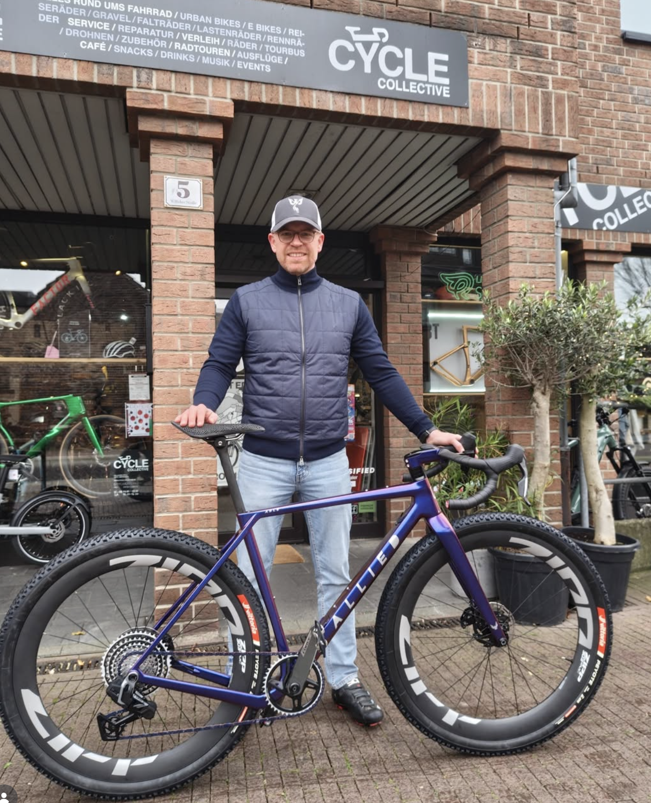 Ein Mann steht vor einem Fahrradgeschäft namens 'Cycle Collective', trägt eine dunkle Weste, eine Kappe und Jeans, und hält ein Rennrad. Das Geschäft hat eine Backsteinfassade mit Fahrrädern im Schaufenster.