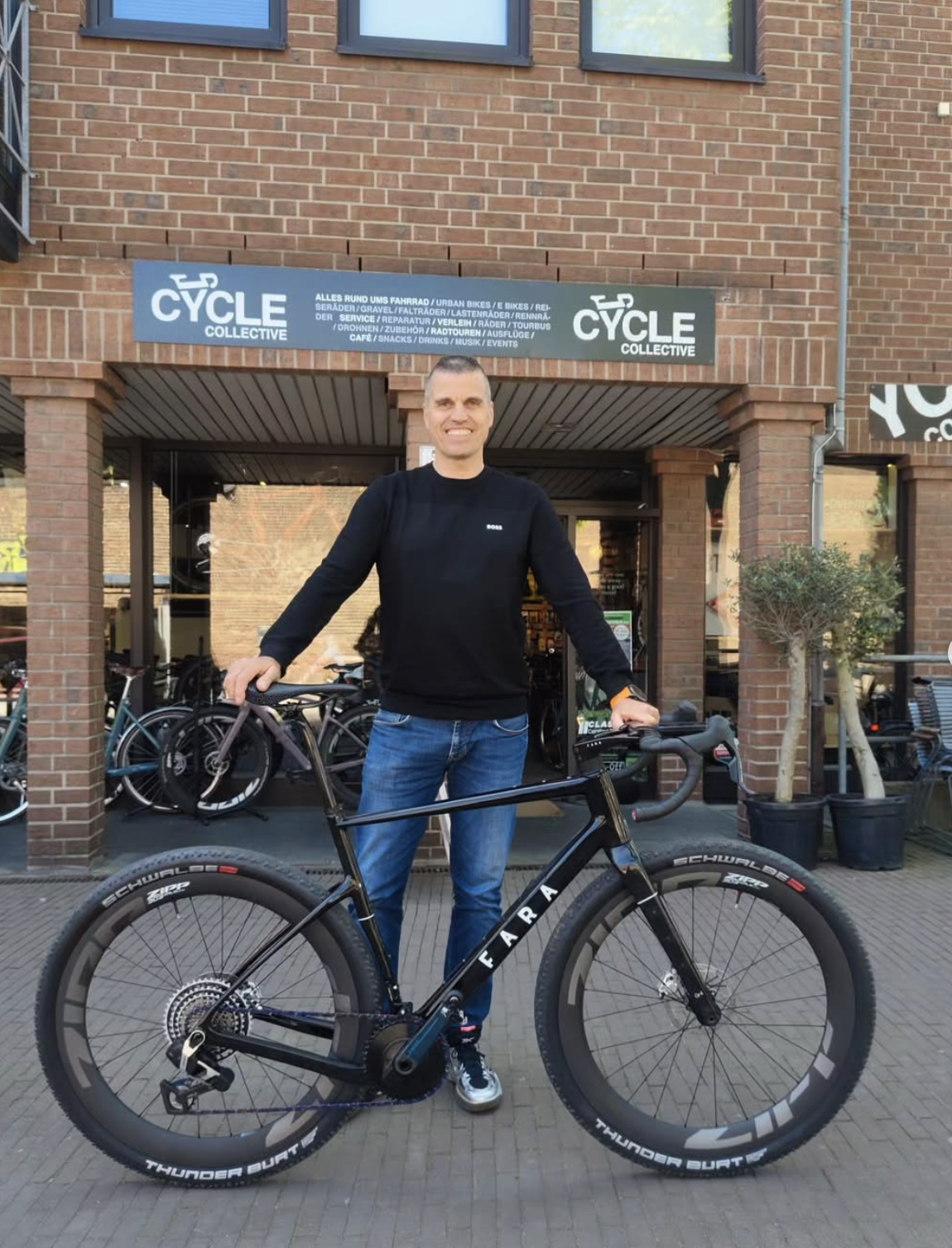 Ein Mann steht vor einem Fahrradladen mit einem schwarzen Fahrrad. Er trägt eine schwarze Jacke und blaue Jeans und lächelt. Im Hintergrund sind Fahrräder und ein Laden mit einem Schild, das auf Fahrrad-Services und -Verleih hinweist.