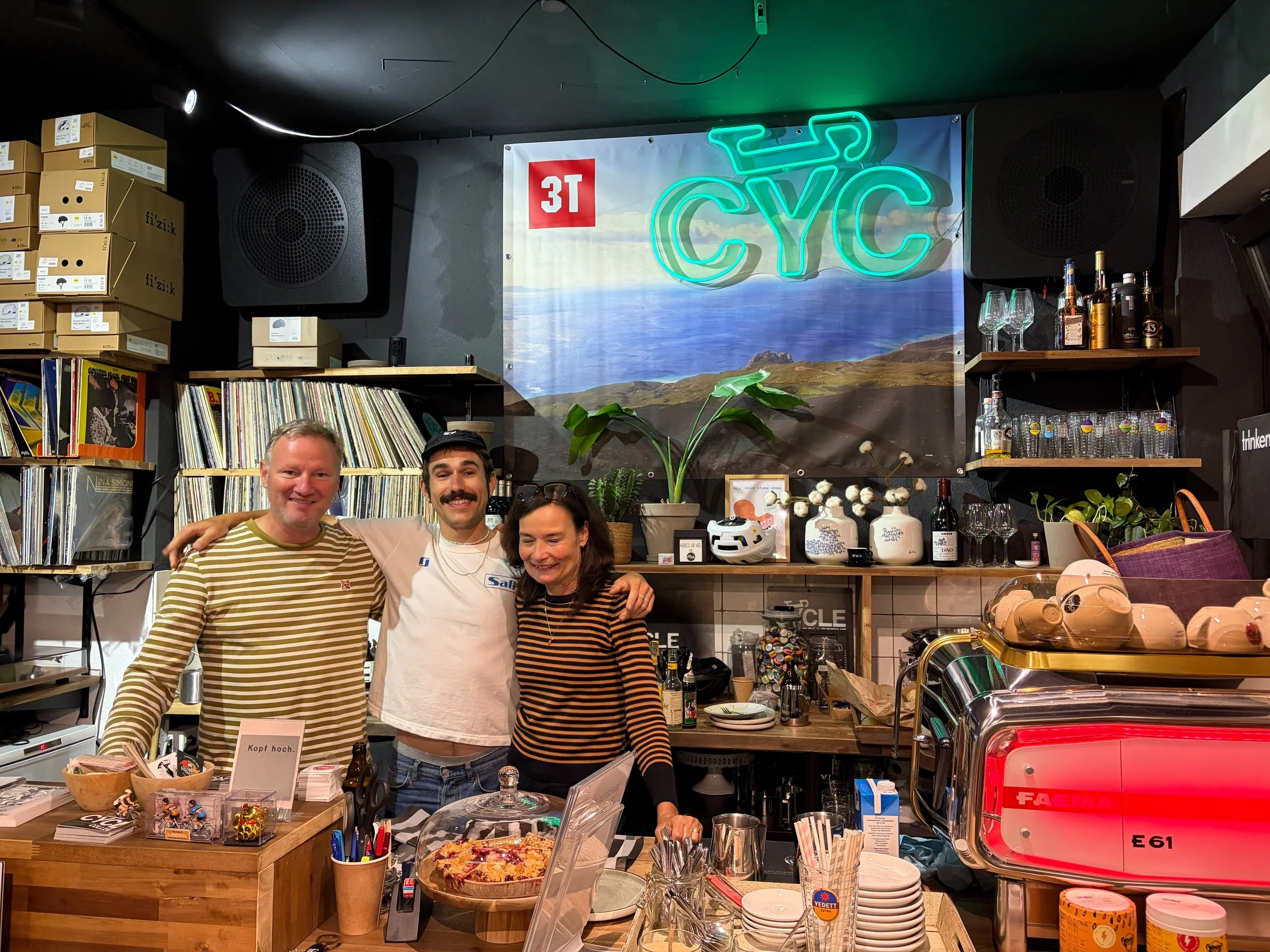 Menschen stehen hinter einer Bar mit Getränken, Gläsern und Deko, im Hintergrund ein neonbeschriftetes Schild mit der Aufschrift 'CYÇ'.