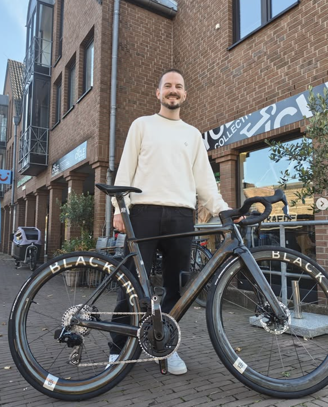 Ein Mann mit dunklen Haaren, Bart und im weißen Sweatshirt steht neben einem schwarzen Rennrad mit 'BLACK', das auf einem Gehweg in einer städtischen Gegend steht. Im Hintergrund sind mehrstöckige Gebäude aus Ziegelsteinen, Geschäfte und Bänke. Es ist Tag und das Wetter ist klar.